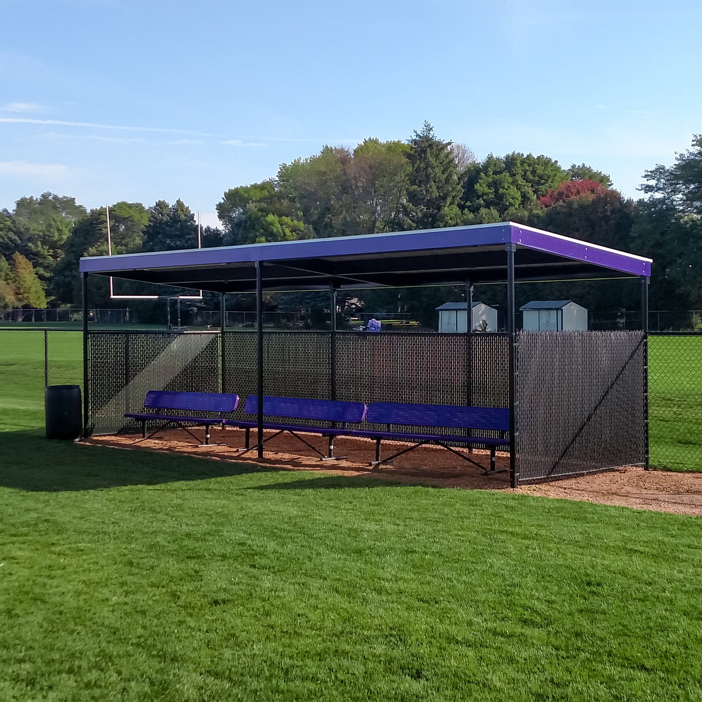 The World Series Dugout features a prefabricated steel roof and benches, set on a grass field and enclosed by a chain-link fence, with trees and sheds in the background beneath a clear sky.