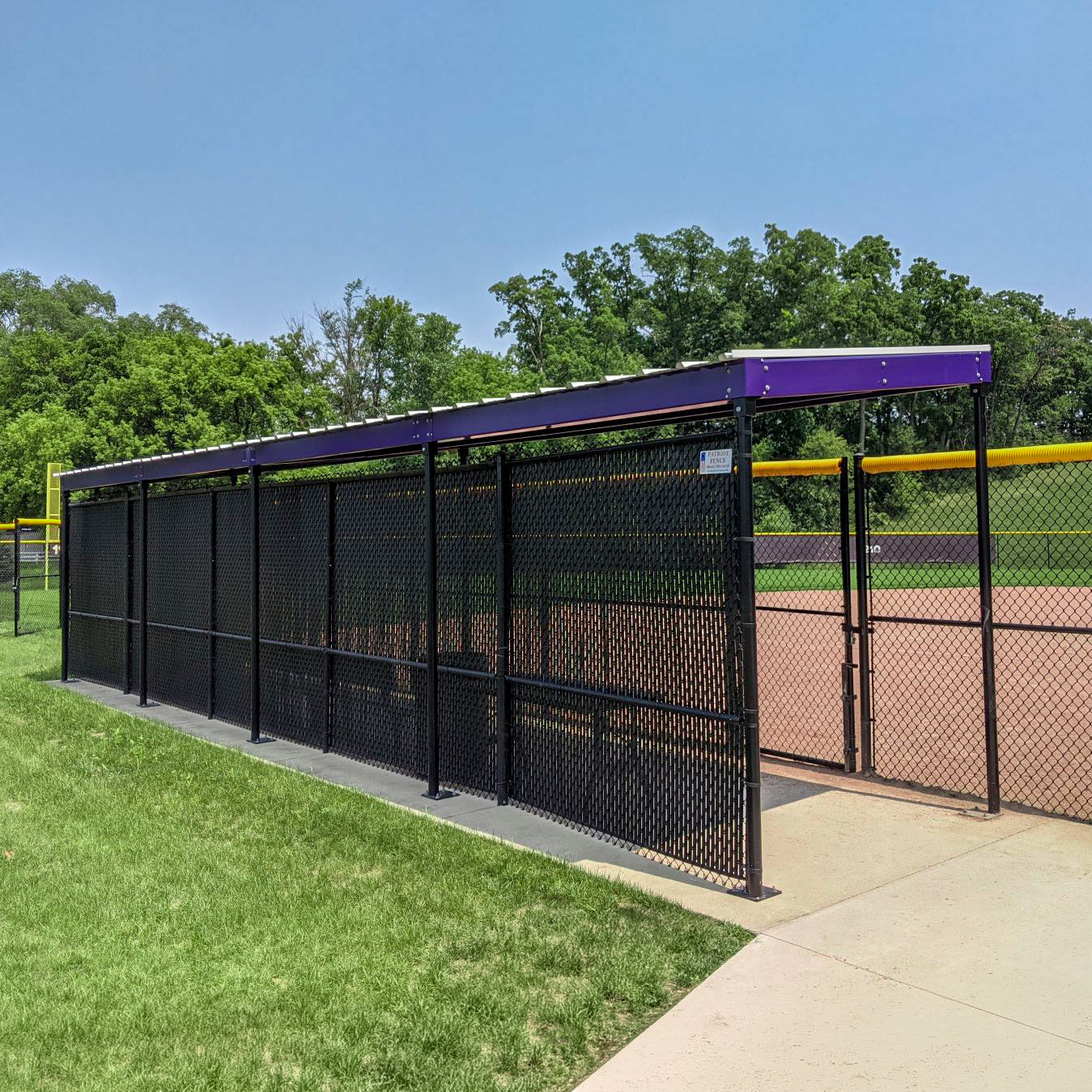 The World Series Dugout features a black chain-link design with a prefabricated steel roof, set beside a baseball field amid green grass and trees—perfect for softball and baseball, and fully wind load compliant.
