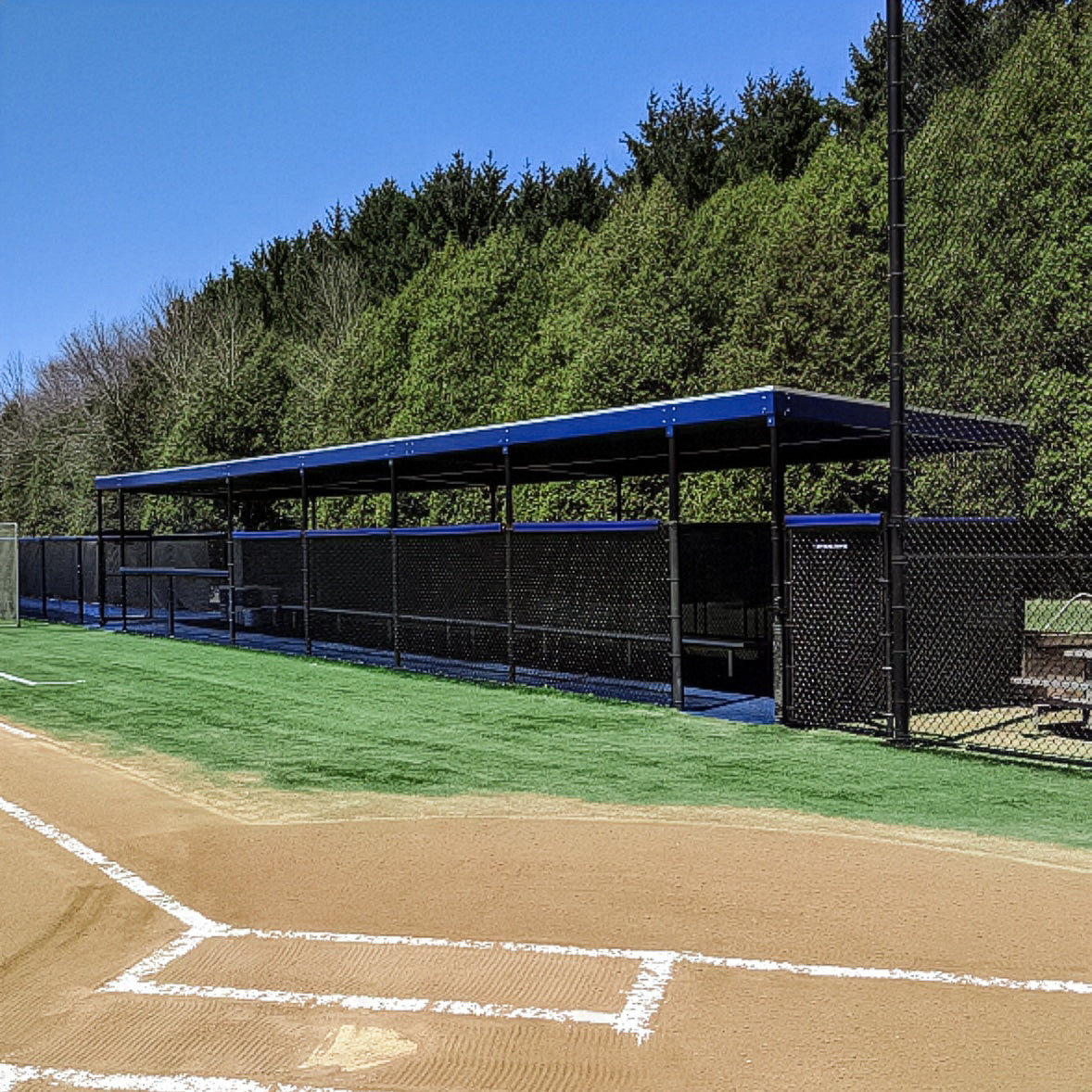 The World Series Dugout, featuring blue trim, benches, and a chain-link fence, sits beside a field with a dirt infield and green turf—a wind load compliant dugout ideal for softball or baseball fields.