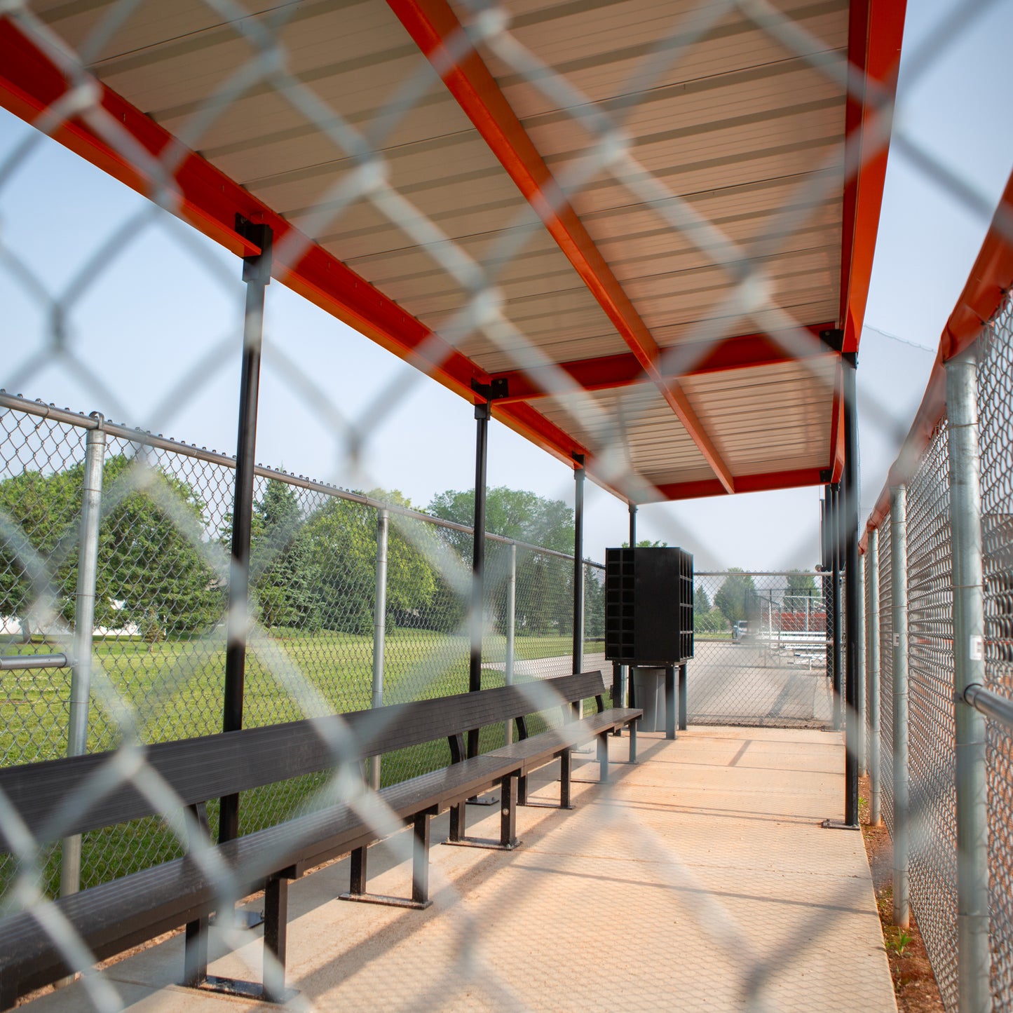 A view through a chain-link fence of an empty World Series Dugout with black benches and a prefabricated steel roof for sun protection, situated beside a grassy field on a sunny day.