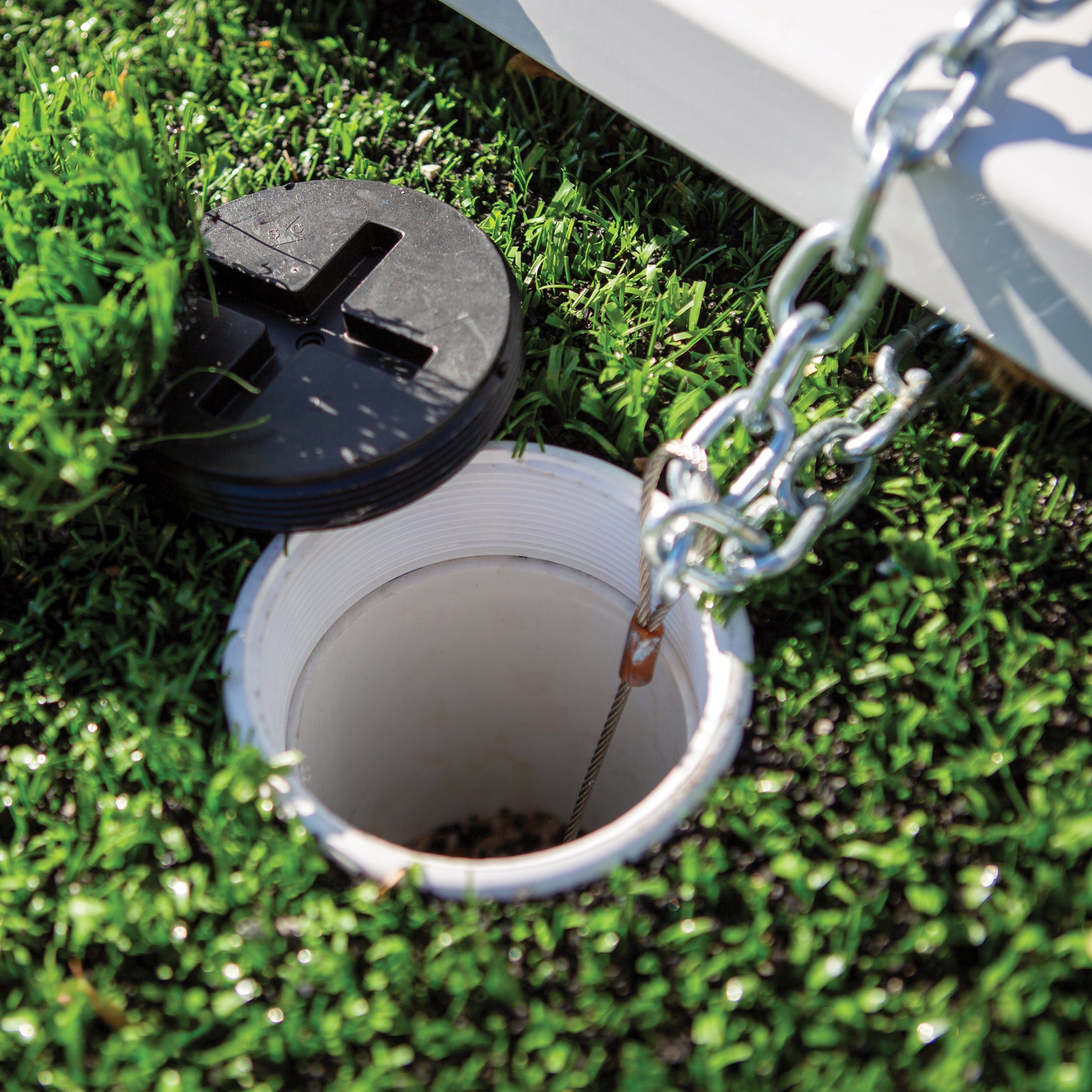 Close-up of an open white drain with a black cap on artificial grass, featuring the Ground Insert For Duckbill Anchor and a metal chain attached nearby.