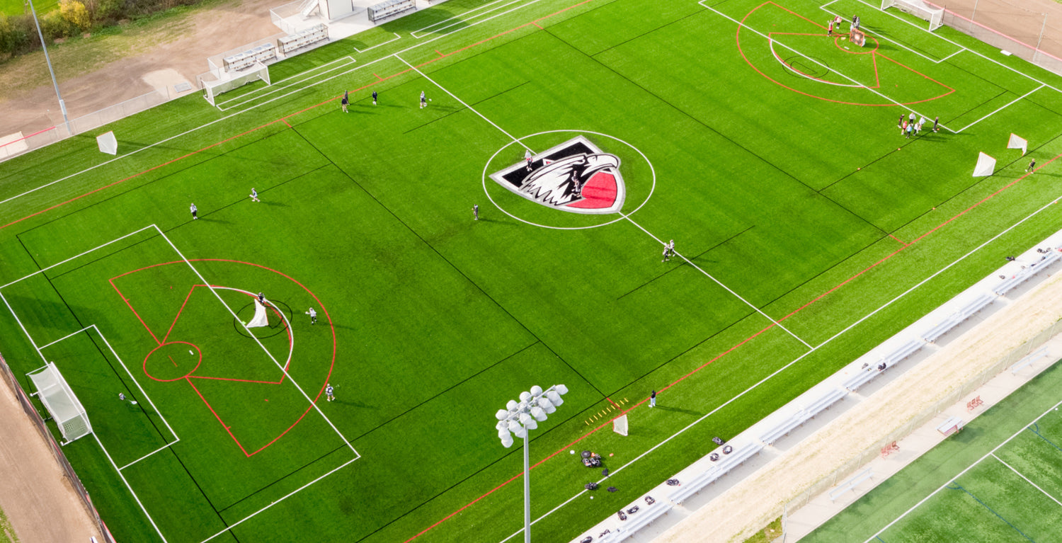 Aerial view of a green artificial turf soccer field with red and white markings, a large wolf logo at the center, and several groups of people playing sports on different sections of the field.