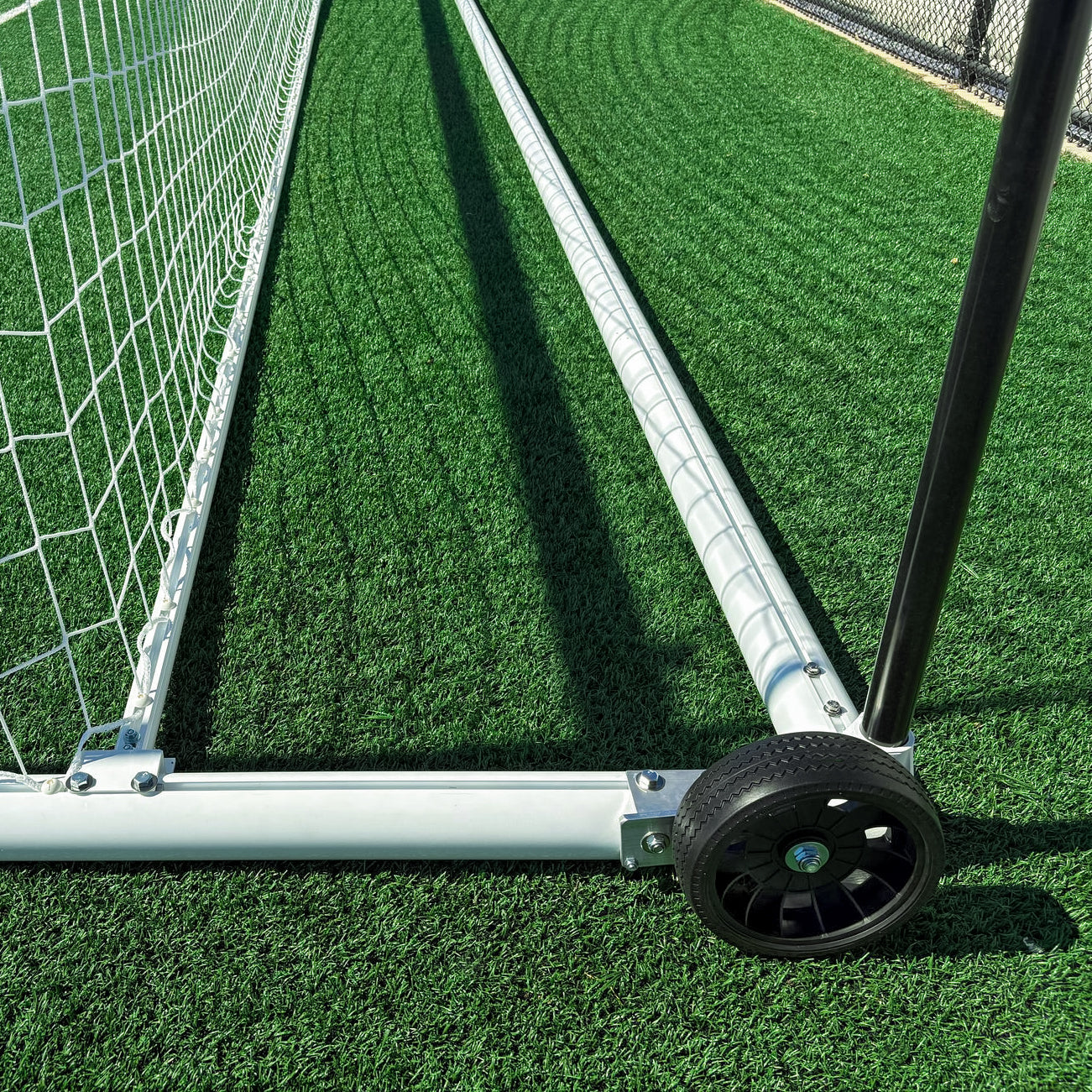 Close-up of the Elite Stadium Soccer Goals base, featuring aluminum stanchion poles and a black wheel on green artificial turf near a chain-link fence. The mobile stanchion design is shown from ground level.
