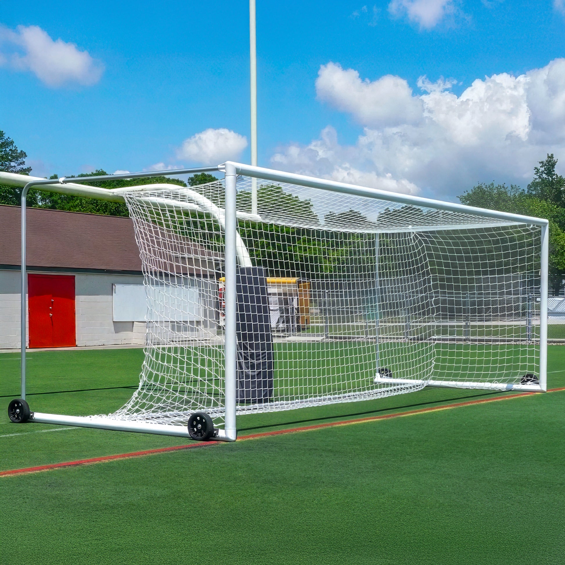 An Elite Box-Style Soccer Goal with a net sits on green artificial turf. This portable soccer goal has wheels for easy movement, with buildings and a blue sky with clouds visible in the background.