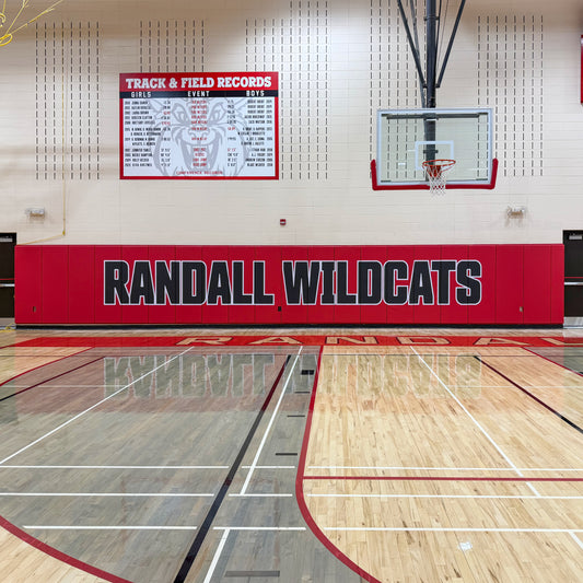 A high school gym features wood floors, a basketball hoop, and red Indoor Protective Padding for walls prominently displaying “RANDALL WILDCATS.” Above, a track & field records board is visible. The clean, brightly lit space showcases the protective padding along the gym walls.