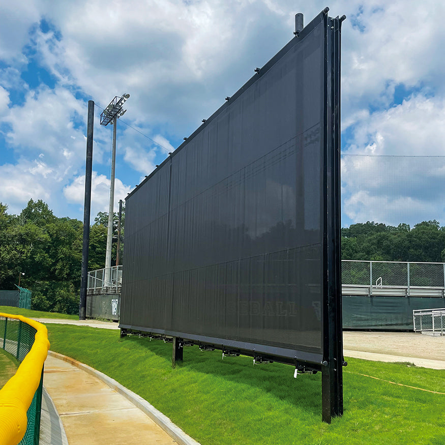 A Jaypro Outfield Batter's Eye (20' x 40') with a large black mesh on a metal frame stands on green grass by a sports field, with trees, fencing, and stadium lights visible under a partly cloudy sky.