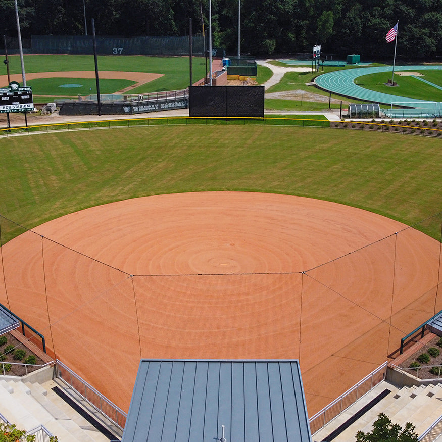 A well-kept baseball field features a dirt infield, lush green outfield, and the Jaypro Outfield Batter's Eye (20' x 40'). Empty bleachers are in front, with a scoreboard, running track, and an American flag visible in the background.