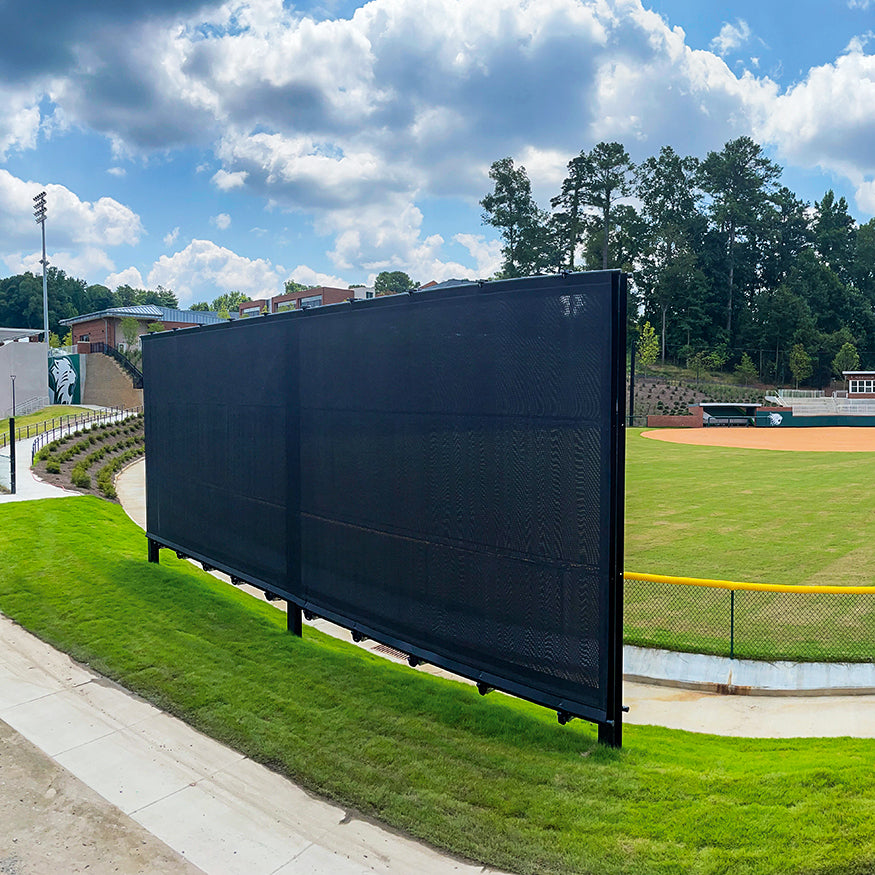 The Jaypro Outfield Batter's Eye (20' x 40') is a large black mesh windscreen along a baseball field fence, with green grass, trees, and a sidewalk under a partly cloudy sky in the background.