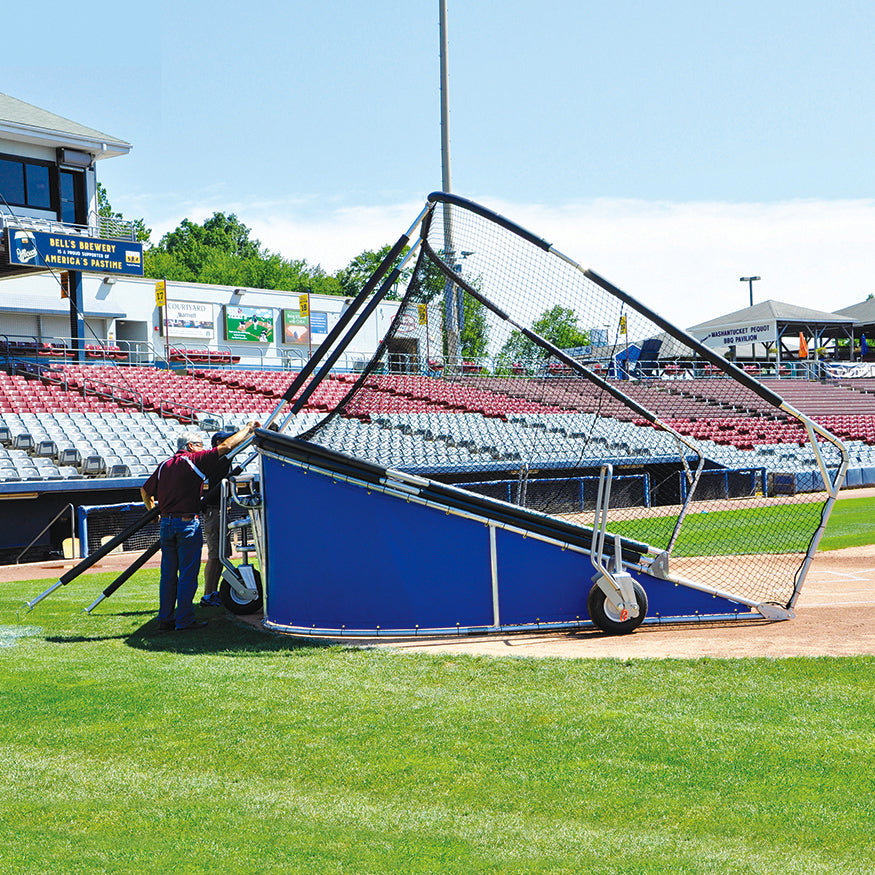Two people stand next to the Jaypro Bomber All-Star Batting Cage | Portable Batting Cage on a baseball field, with empty stadium seating and buildings in the background under a clear sky.