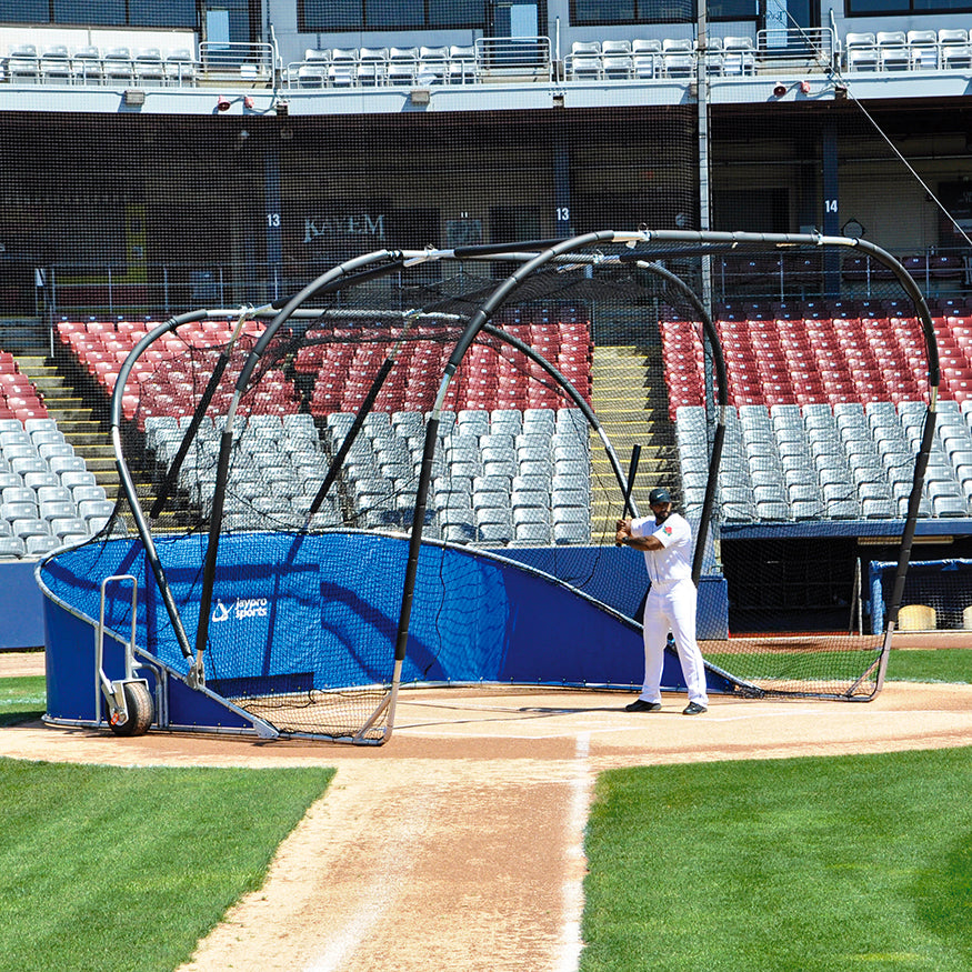 A baseball player gets ready to bat inside the Jaypro Bomber All-Star Portable Batting Cage on the field, with empty stadium seats in the background.