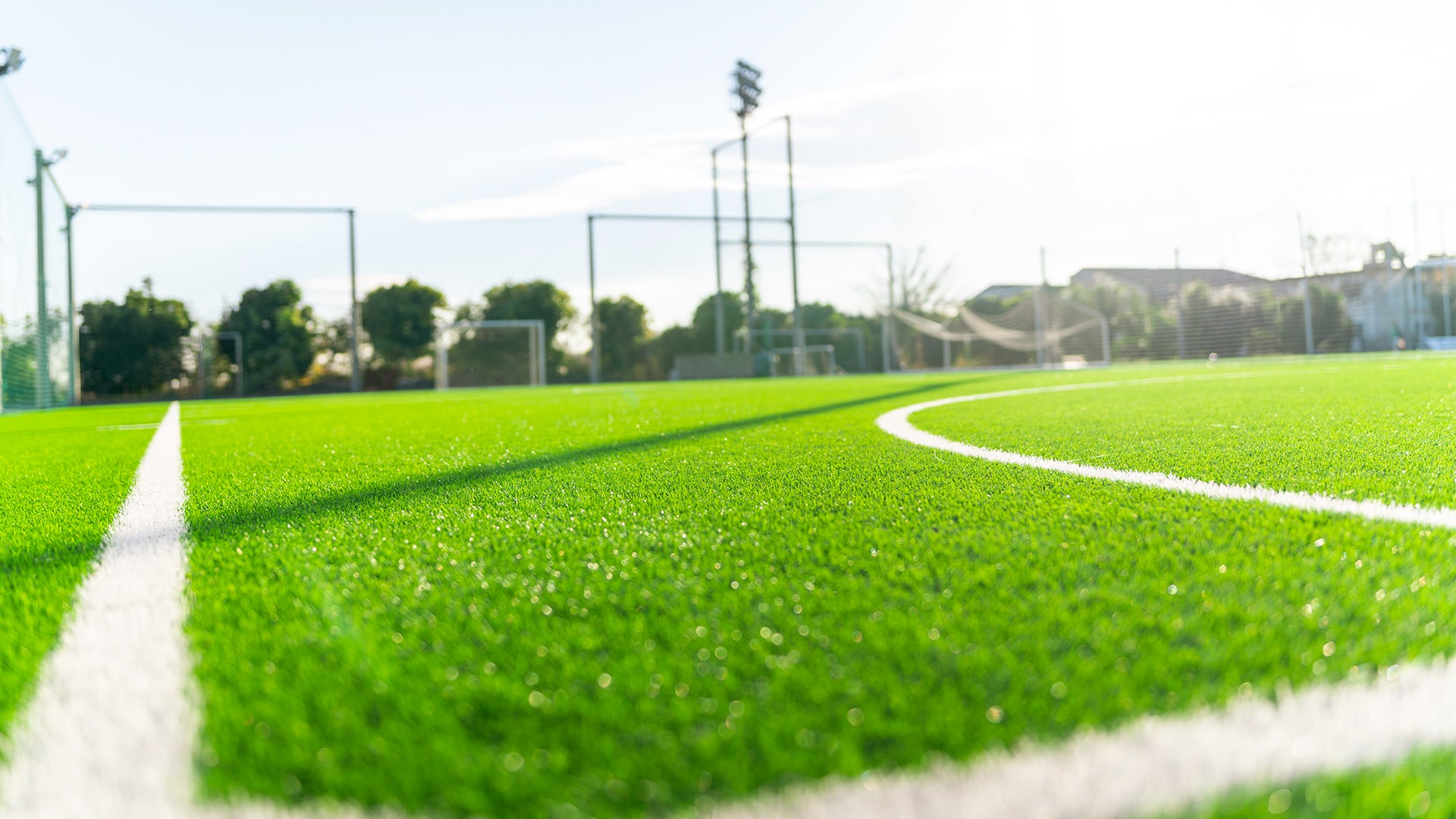 A close-up view of a vibrant green artificial turf soccer field with bright white boundary lines, photographed on a sunny day with trees, goal posts, and floodlights in the background.