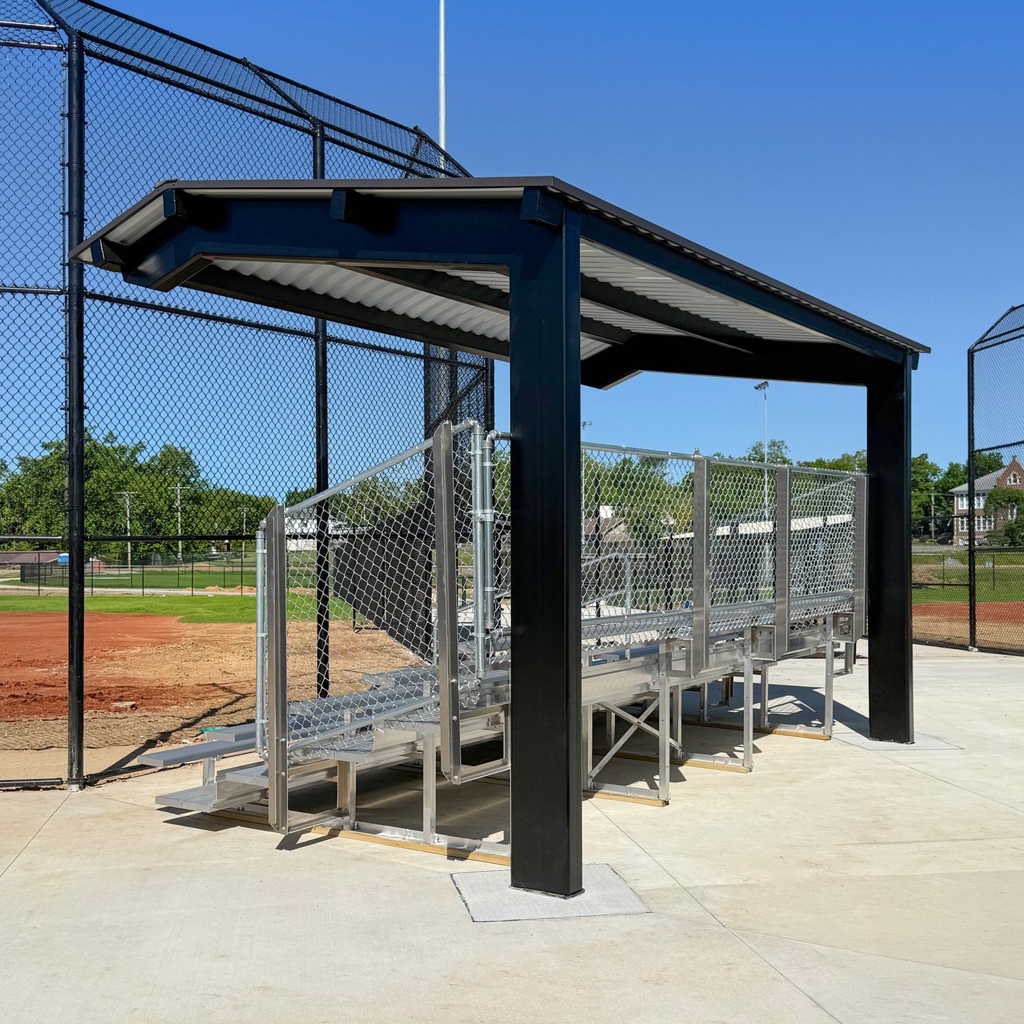 Bleachers with a Premium Hard Shell Bleacher Cover and chain-link fencing sit beside a baseball field, bordered by black fence, with trees and buildings in the background on a sunny day.