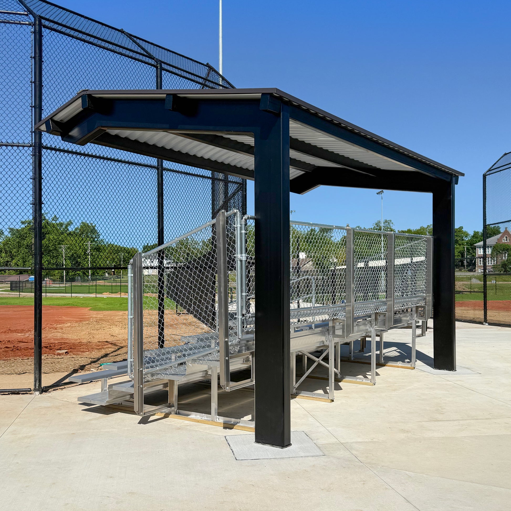 Bleachers with a Premium Hard Shell Bleacher Cover and chain-link fencing sit beside a baseball field, bordered by black fence, with trees and buildings in the background on a sunny day.