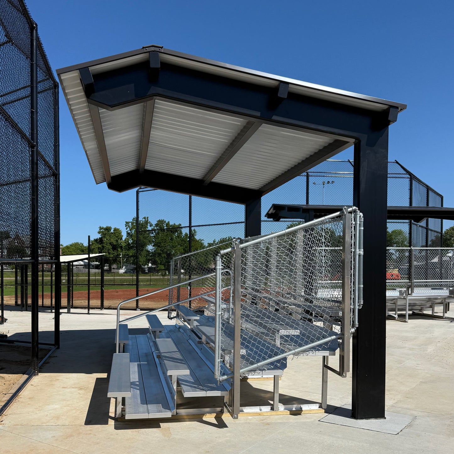 The Premium Hard Shell Bleacher Cover provides shaded seating over metal bleachers with chain-link fencing, installed on concrete at a baseball field, ensuring ample spectator comfort beneath blue skies and surrounding trees.