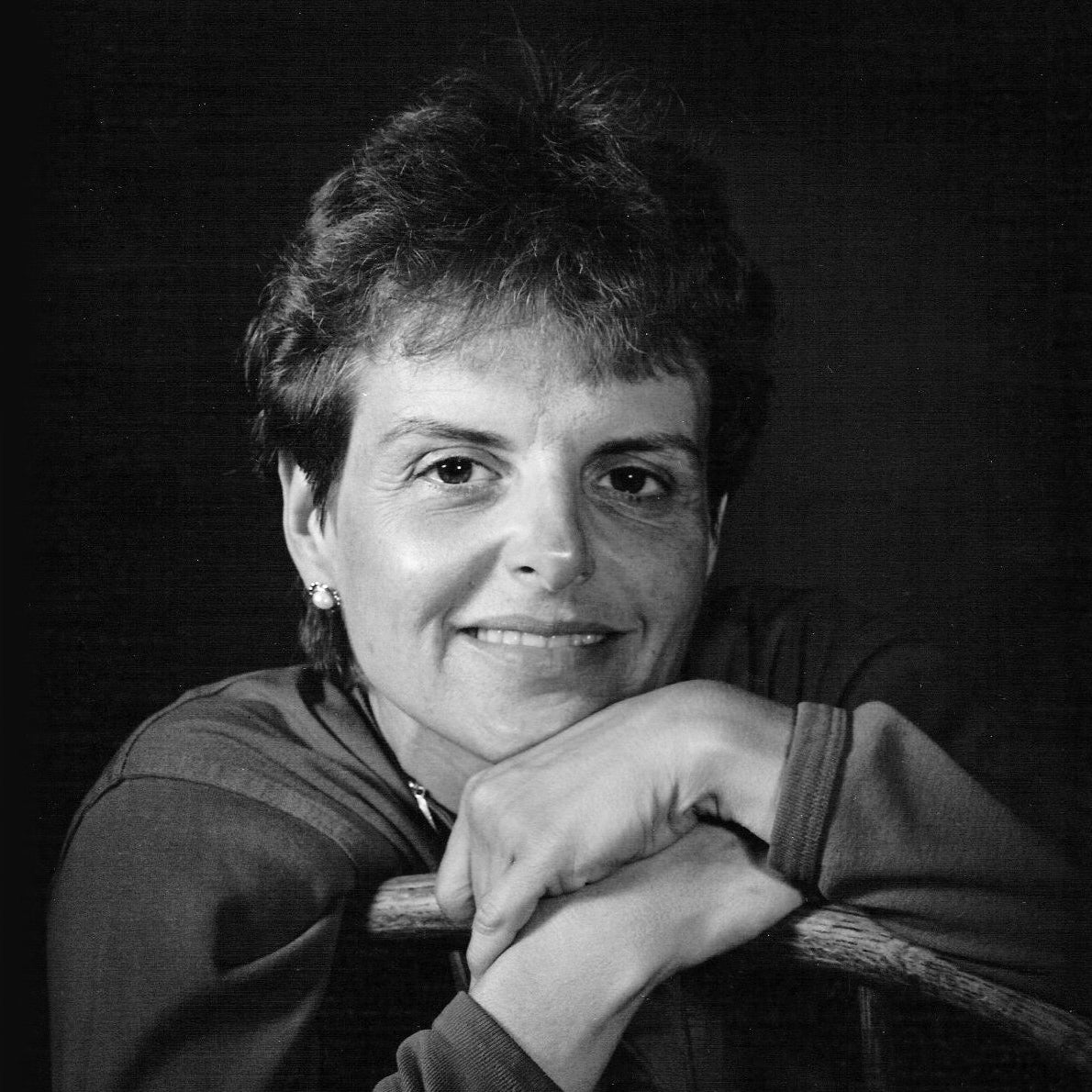 Black and white portrait of a person with short hair, smiling gently and resting their chin on their hands, which are folded over the back of a wooden chair.