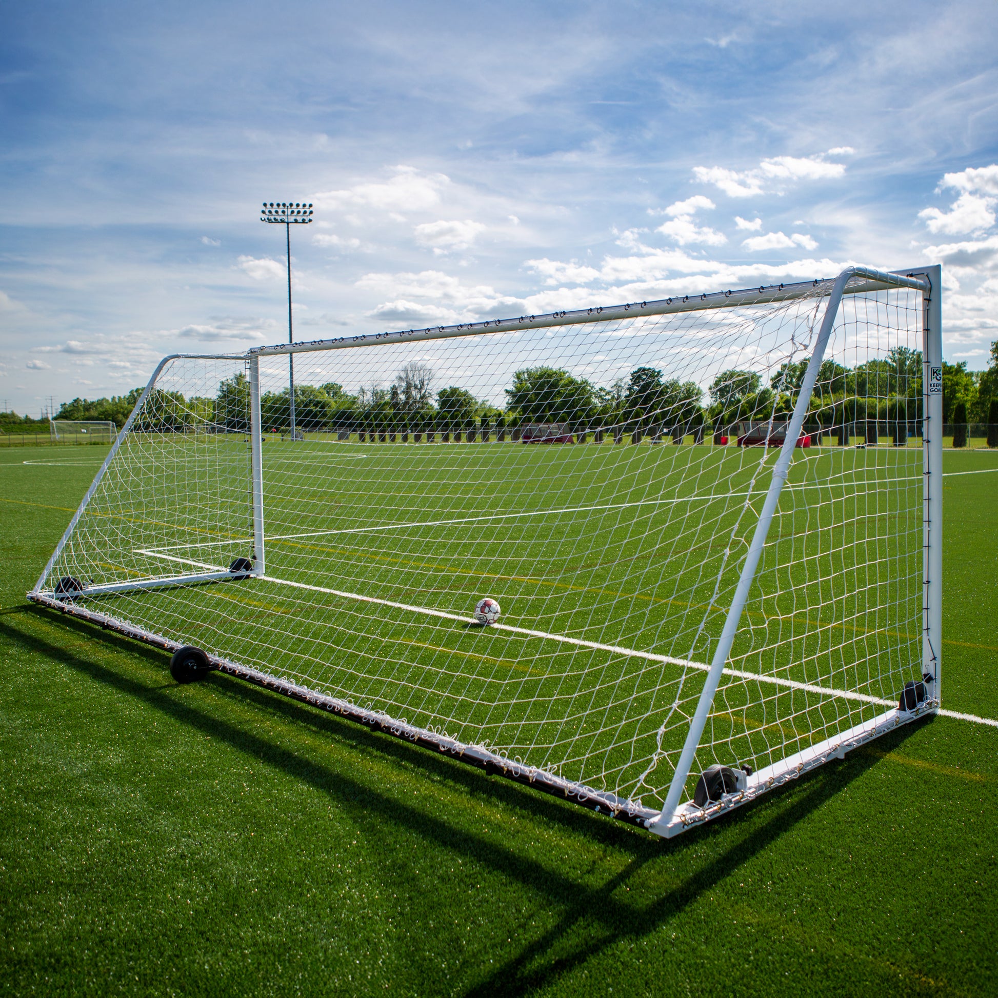 The Ultimate Wheeled Premier Soccer Goals stand on green artificial turf beneath a blue sky with scattered clouds. A soccer ball sits inside the goal, with trees and a light pole visible in the background.