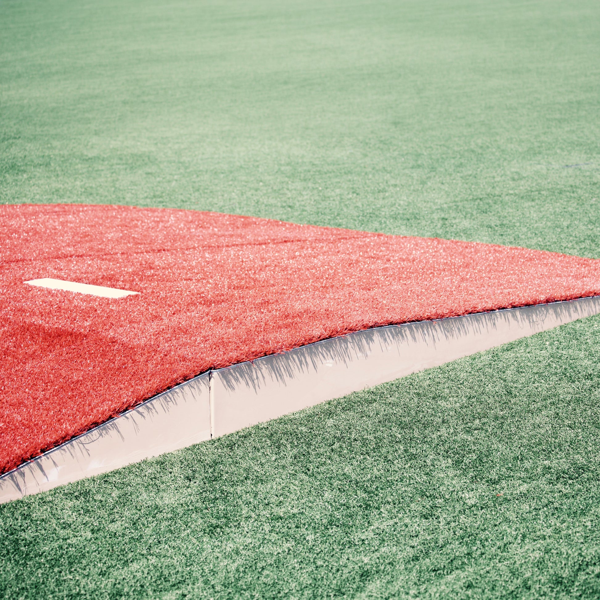 A close-up of The Perfect Mound Portable Bullpen Pitching Mound Series in red on a green artificial field, featuring visible white seams along the edge for both on field and off field configurations.