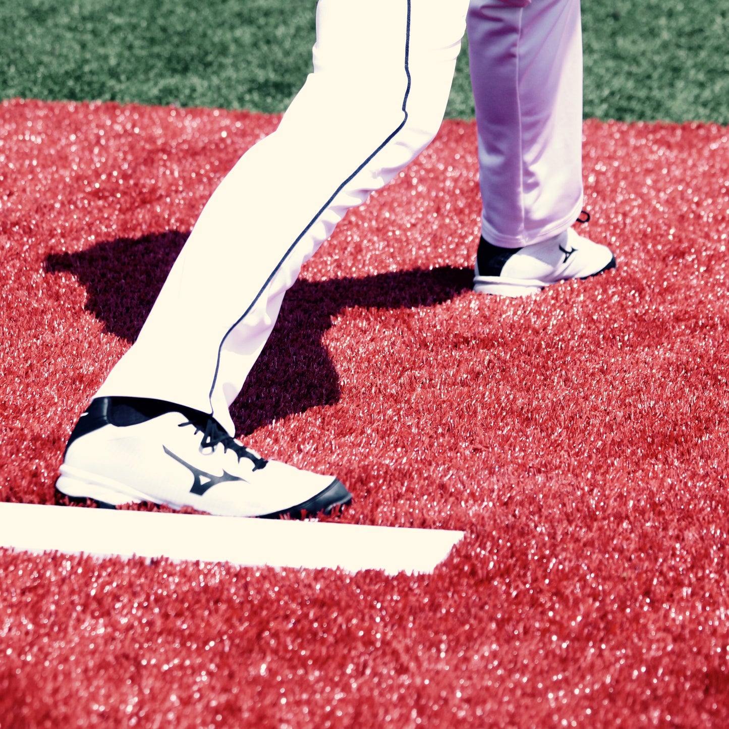 A close-up of a baseball player’s legs in white pants and black-and-white cleats stands on bright red turf near a white foul line, ready on The Perfect Mound Portable Bullpen Pitching Mound Series.