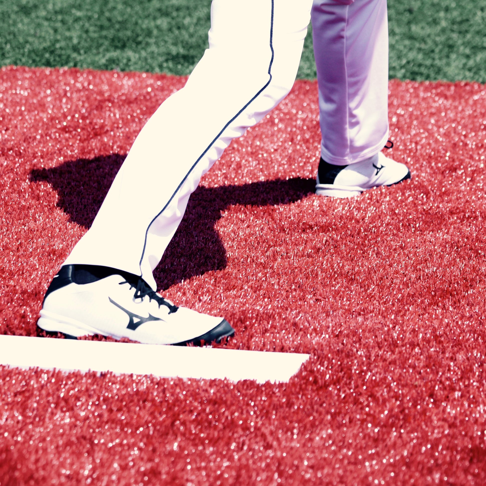 A close-up of a baseball player’s legs in white pants and black-and-white cleats stands on bright red turf near a white foul line, ready on The Perfect Mound Portable Bullpen Pitching Mound Series.