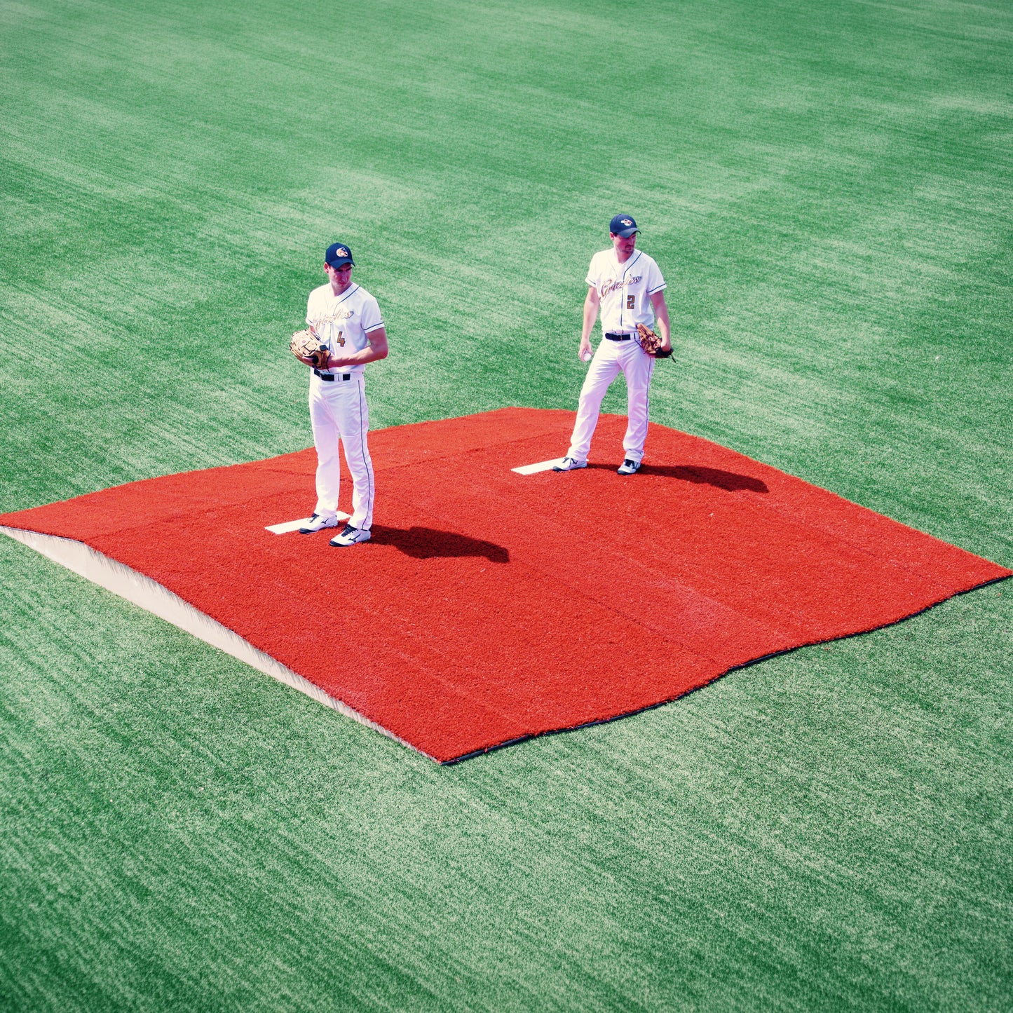 Two baseball players in white uniforms stand on adjacent The Perfect Mound Portable Bullpen Pitching Mound Series on a green field, each holding a glove and ball, facing forward.