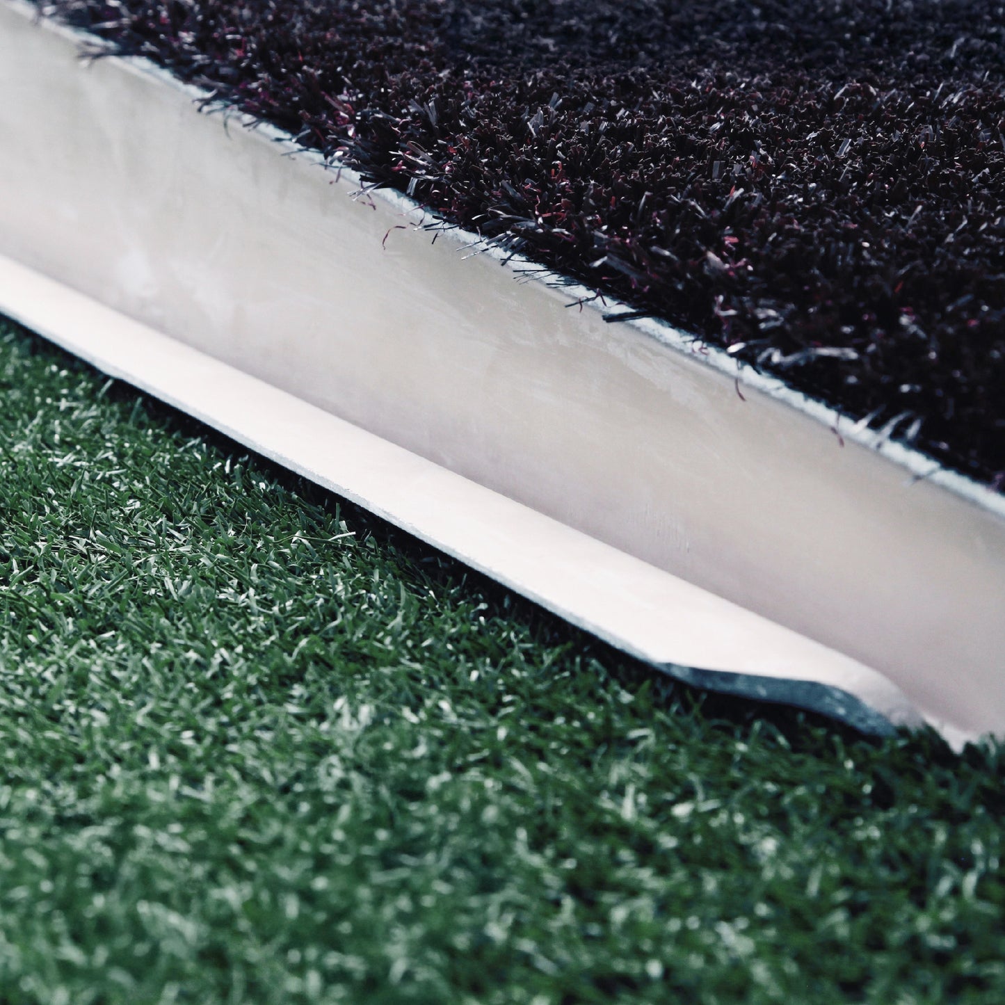 Close-up of The Perfect Mound Adult Portable Pitching Mound’s turf edge shows a cross-section with black infill and green synthetic grass separated by a white barrier—designed to meet MLB specs for portable pitching.