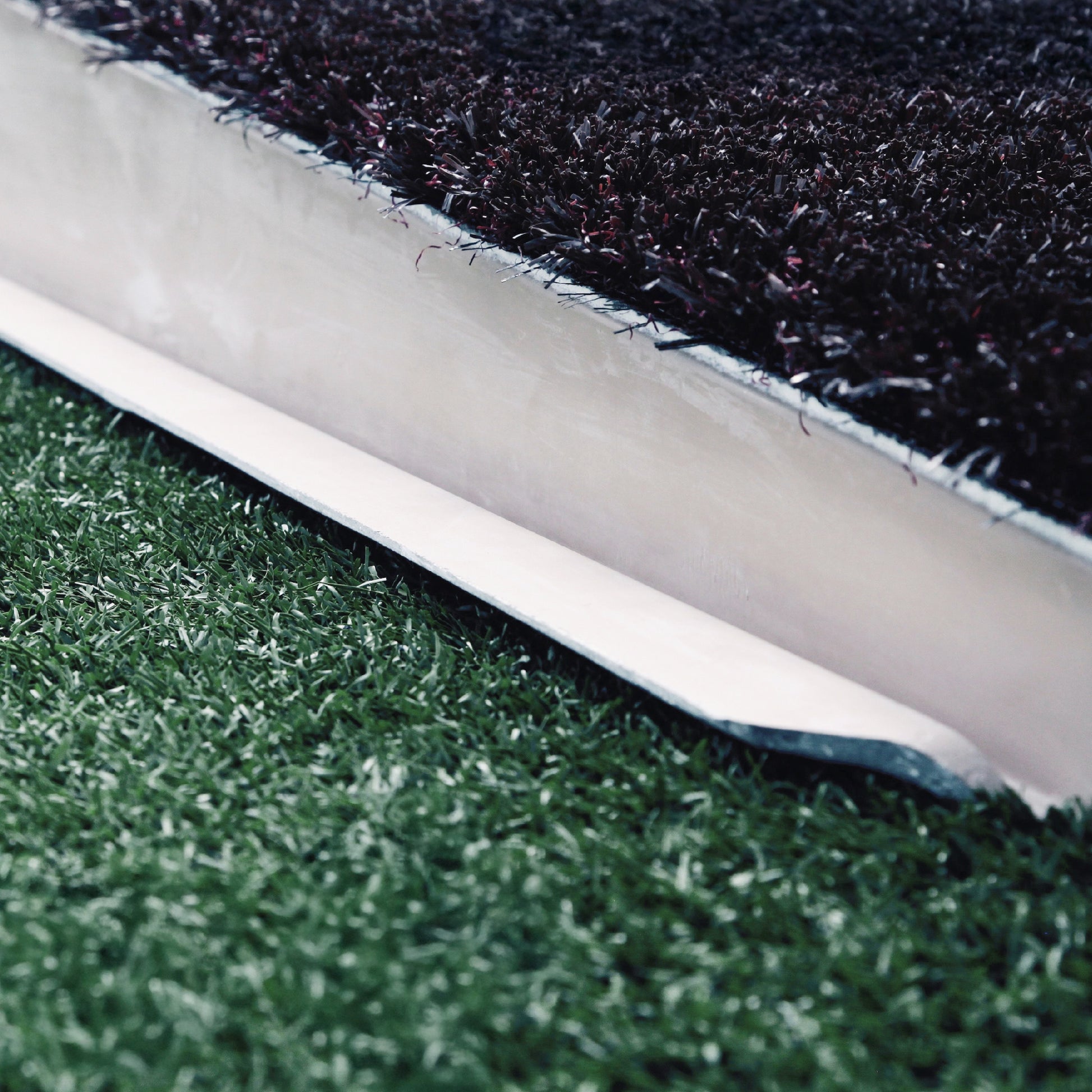 Close-up of The Perfect Mound Adult Portable Pitching Mound’s turf edge shows a cross-section with black infill and green synthetic grass separated by a white barrier—designed to meet MLB specs for portable pitching.