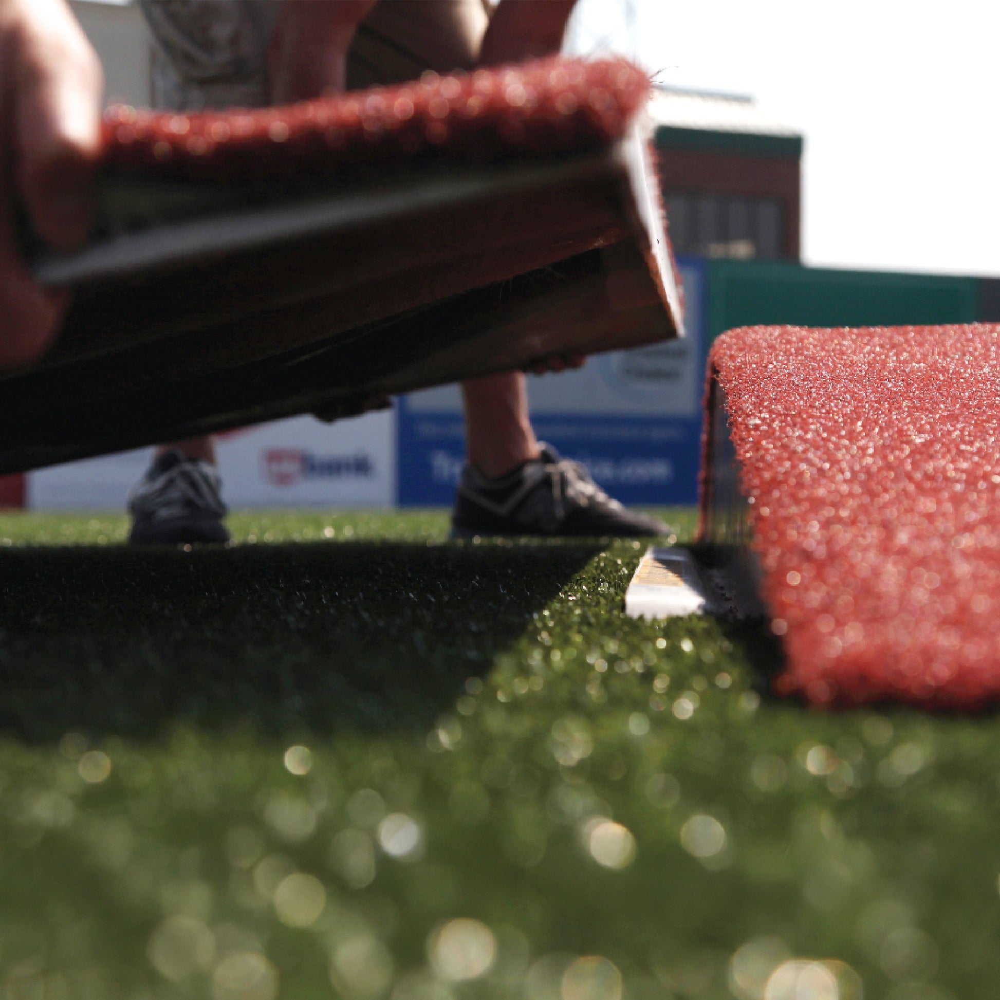 A person installs red artificial turf on a green field, aligning it near The Perfect Mound Youth Portable Pitching Mound for Little League & Youth Baseball. Their hands and feet are visible, with blurred buildings and a sign in the background.
