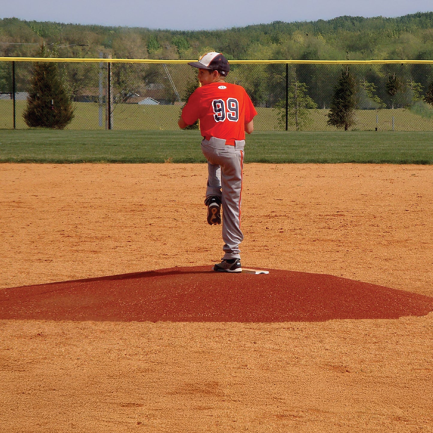 A young baseball player in a red #99 jersey prepares to pitch on The Perfect Mound Youth Portable Pitching Mound at an outdoor field surrounded by trees.