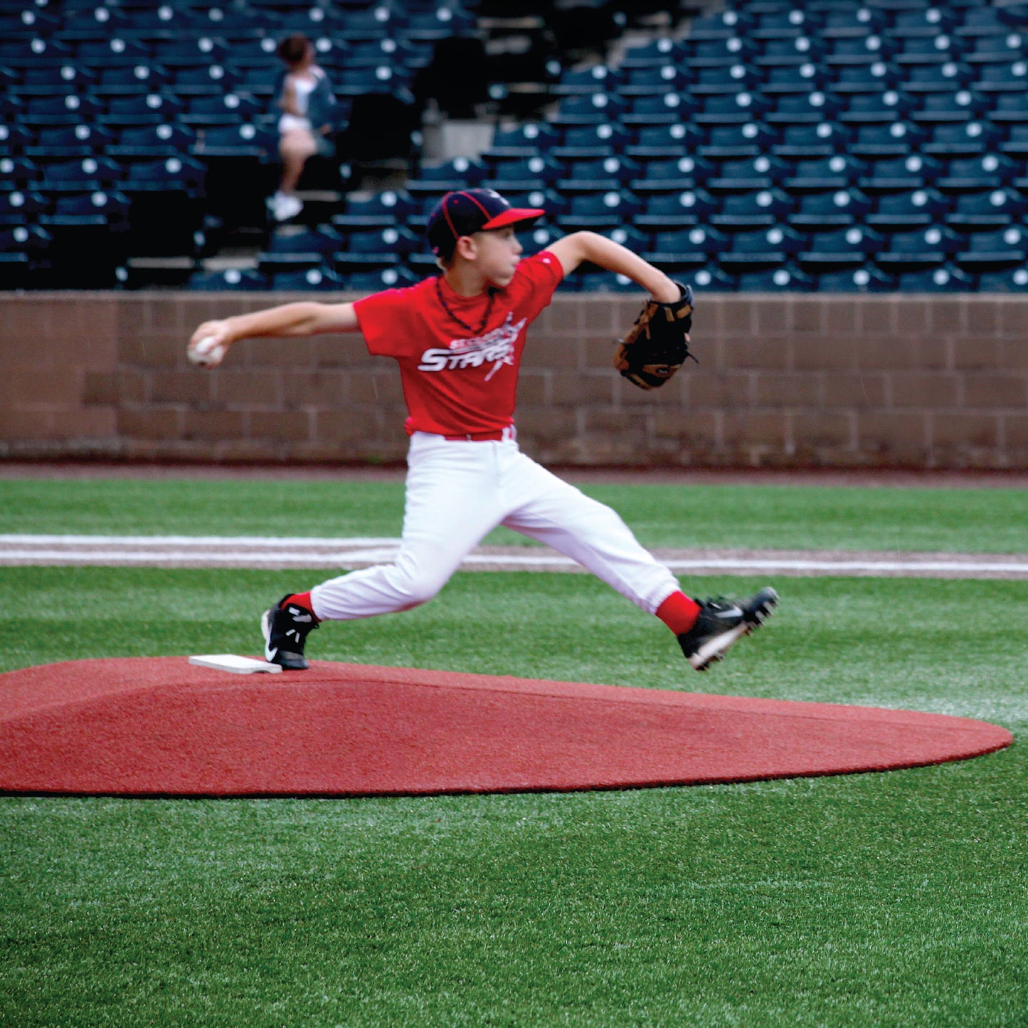A young baseball player in a red uniform pitches mid-throw on The Perfect Mound Youth Portable Pitching Mound, designed for Little League & youth baseball, with empty blue stadium seats in the background.