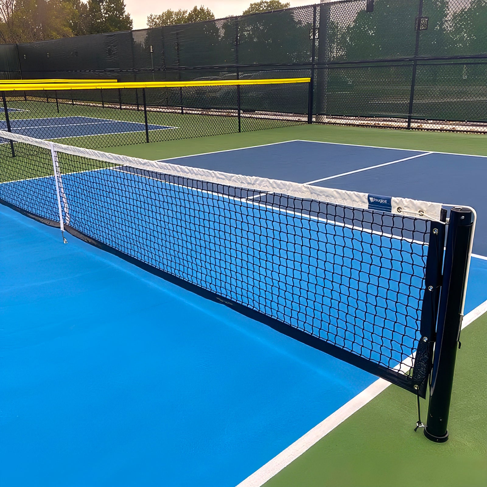 A close-up of a blue and green outdoor pickleball court featuring the Short Pickleball Net | 21'9" L x 31" H Tournament Net, set at regulation height, with a chain-link fence and trees in the background.