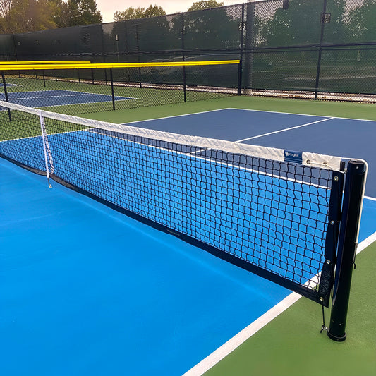 A close-up of a pickleball court featuring the Short Pickleball Net (31" H x 21'9" L) with crisp white lines on a blue and green surface; a chain-link fence and trees can be seen in the background.