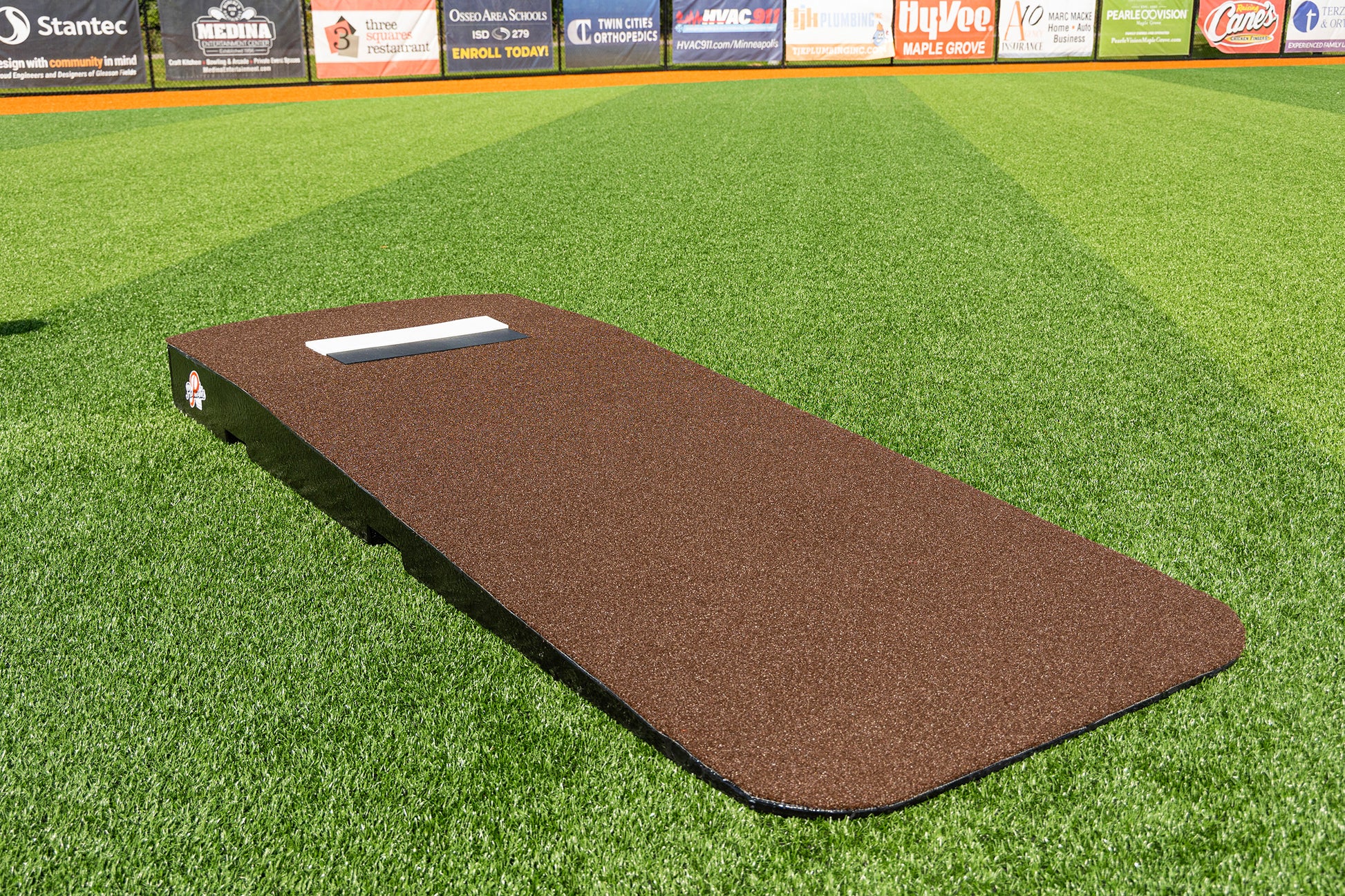 The Portolite Standard One-Piece Practice Mound | Durable Mound for All Ages, featuring a white pitching rubber, is positioned on an artificial turf baseball field with advertising banners visible on the outfield fence in the background.
