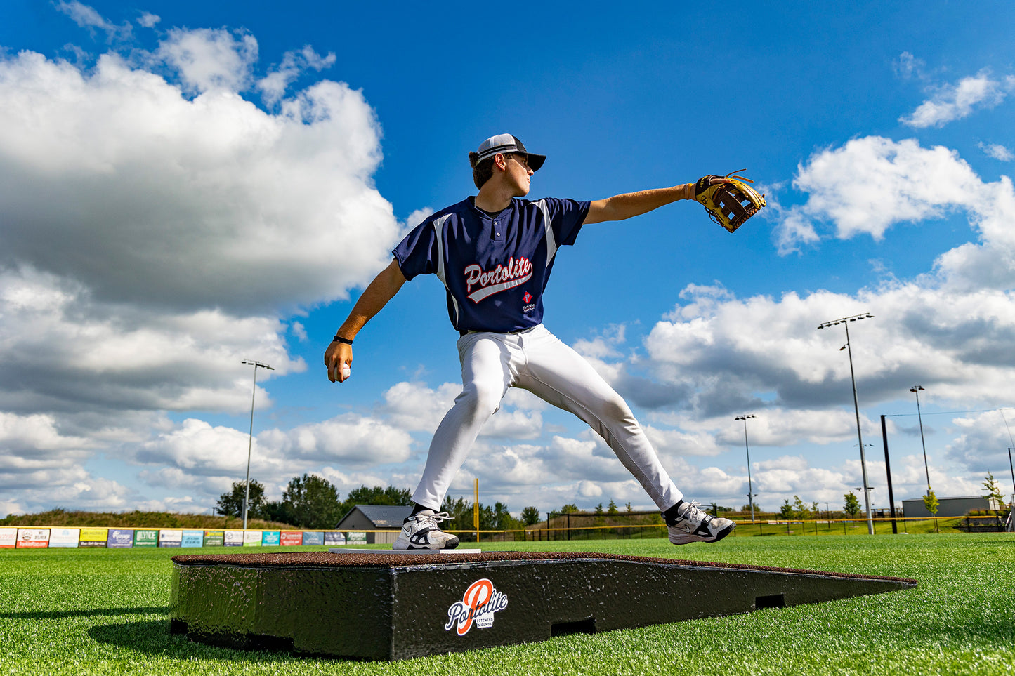 A baseball player in a navy jersey and white pants stands mid-pitch on the Portolite Standard One-Piece Practice Mound—a durable mound for all ages—glove raised, on a sunny, partially cloudy day at an outdoor field.