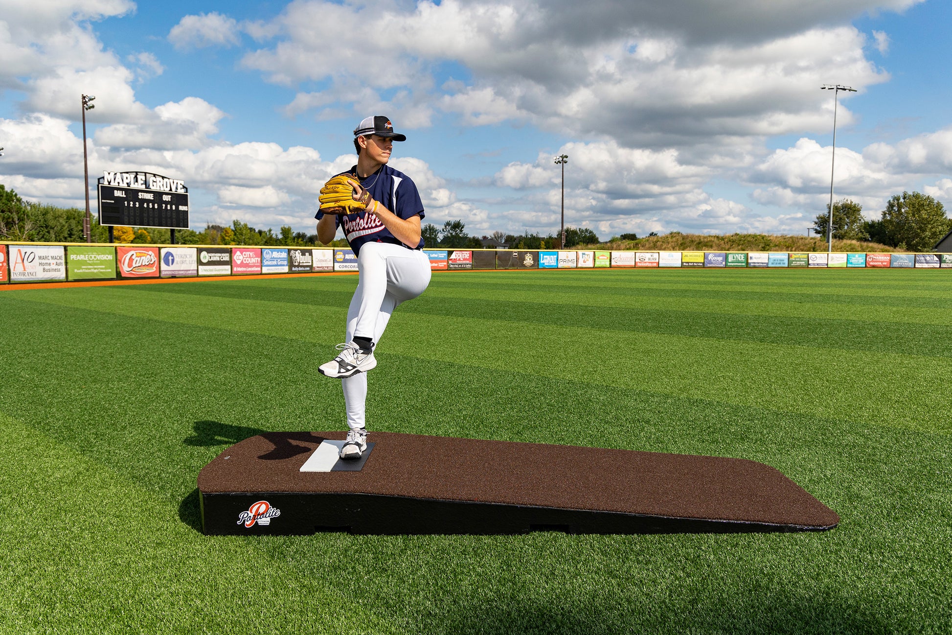 A baseball pitcher in wind-up stance atop the Portolite Standard One-Piece Practice Mound | Durable Mound for All Ages, set on a turf field at Maple Grove baseball stadium under a partly cloudy sky.