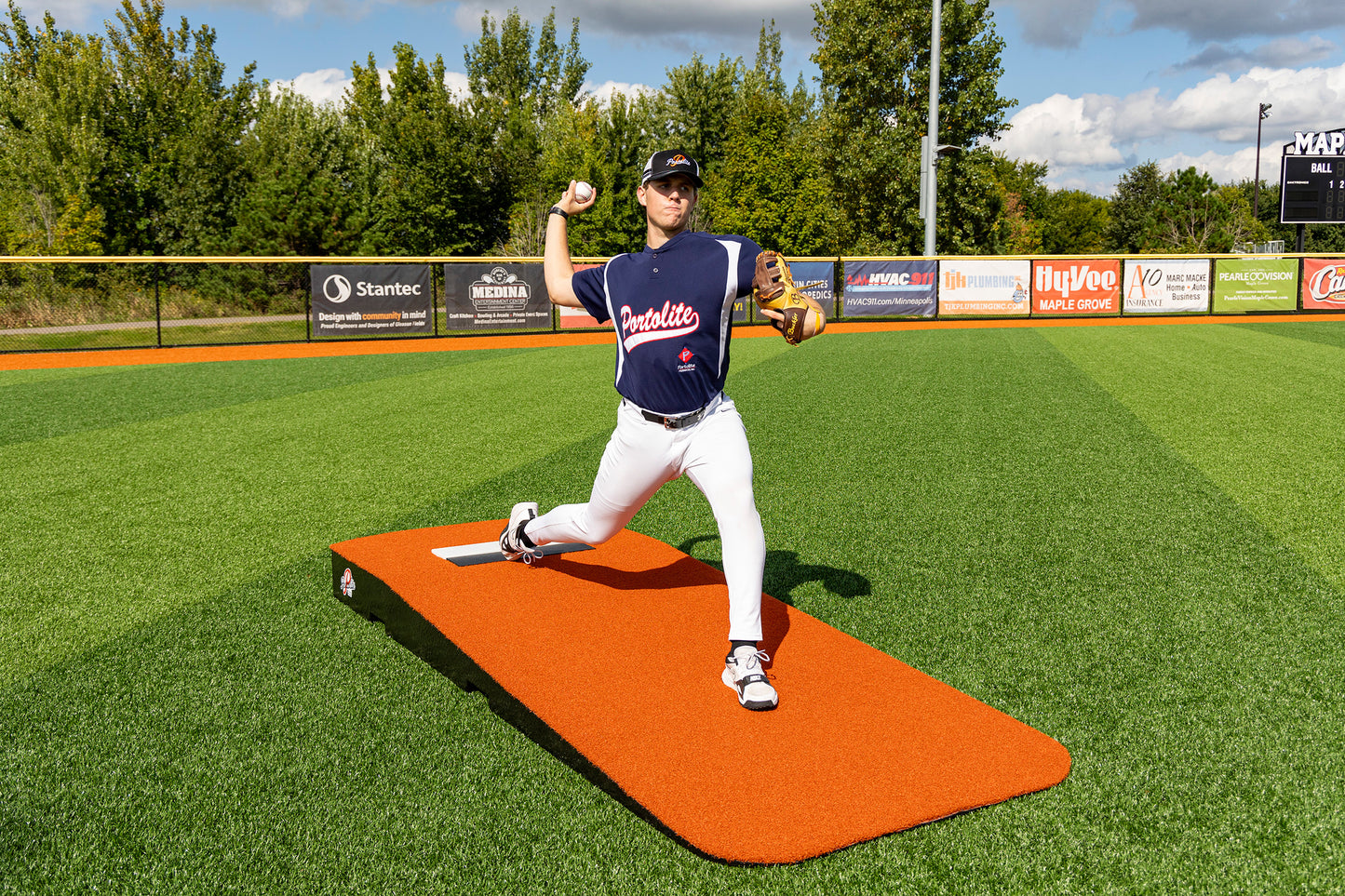 A baseball player in a blue jersey and white pants pitches from the Portolite Standard One-Piece Practice Mound, a durable mound for all ages, set on a bright orange mound on a green field, surrounded by trees and advertising banners.