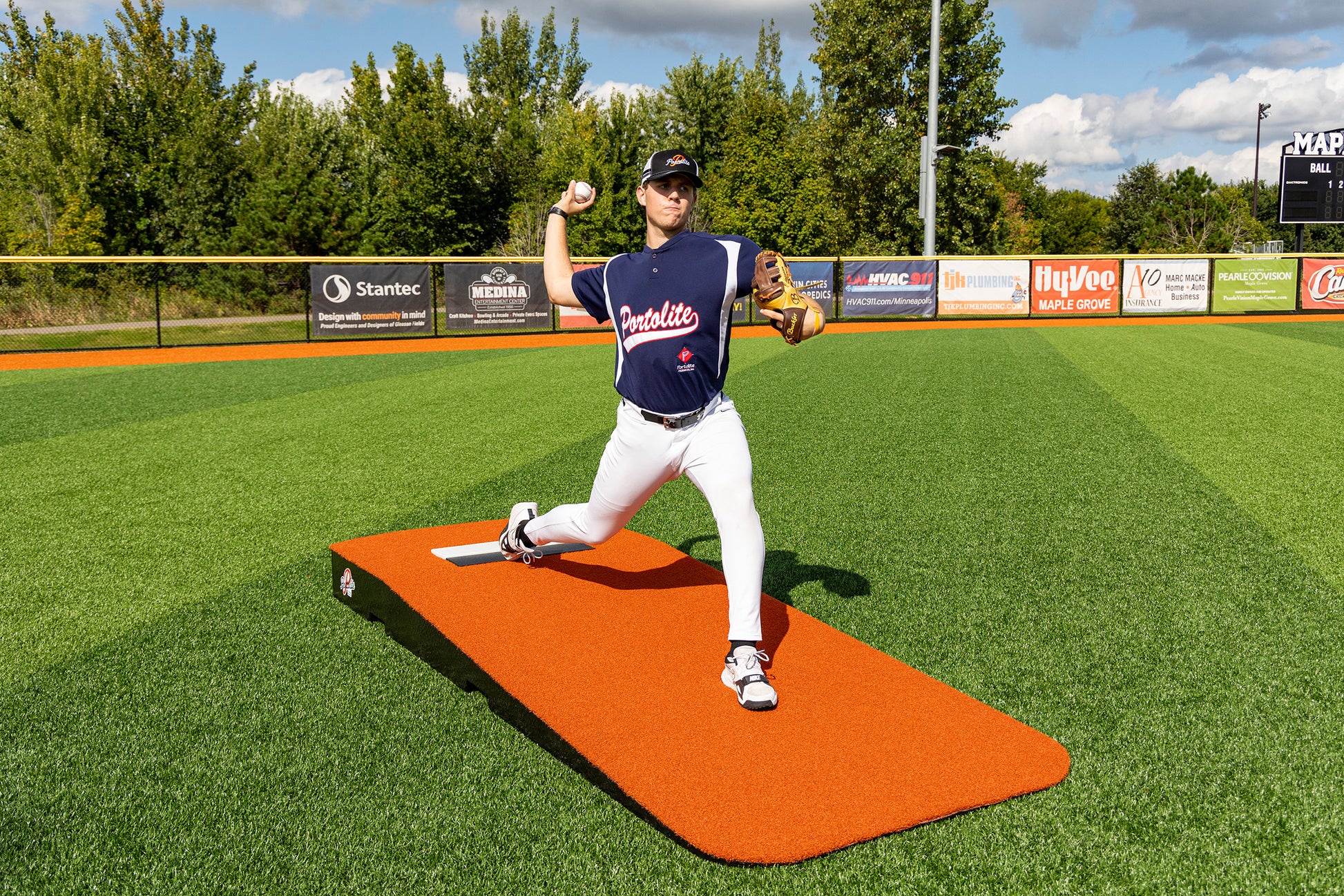 A baseball player in a blue jersey and white pants pitches from the Portolite Standard One-Piece Practice Mound, a durable mound for all ages, set on a bright orange mound on a green field, surrounded by trees and advertising banners.