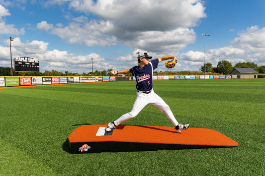 A baseball player in a navy uniform practices pitching on the Portolite Standard One-Piece Practice Mound—a durable mound for all ages—at a sunny outdoor stadium, with a scoreboard and advertisements visible in the background.