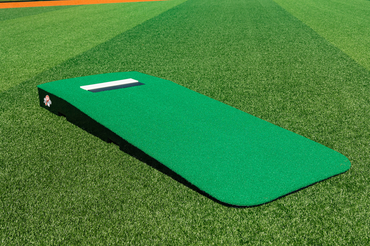 The Portolite Standard One-Piece Practice Mound, a durable mound designed for all ages, sits on the baseball field’s grass with a white rubber strip at the top and is surrounded by striped mowing patterns.