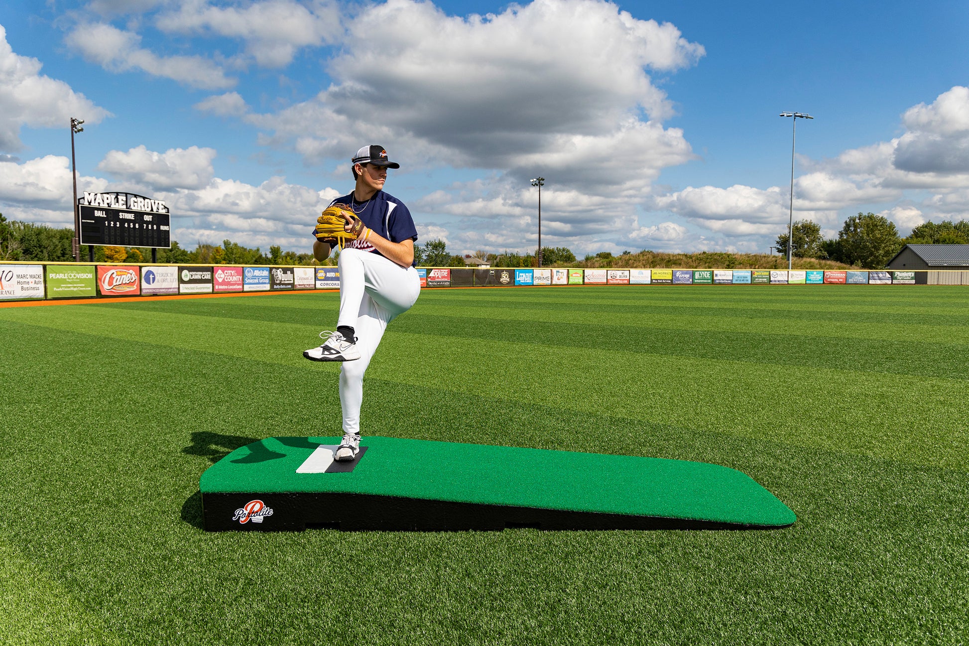 A baseball player in uniform stands on the Portolite Standard One-Piece Practice Mound—a durable mound for all ages—positioned on a green field, preparing to pitch under a partly cloudy sky with a scoreboard and advertisements visible in the background.