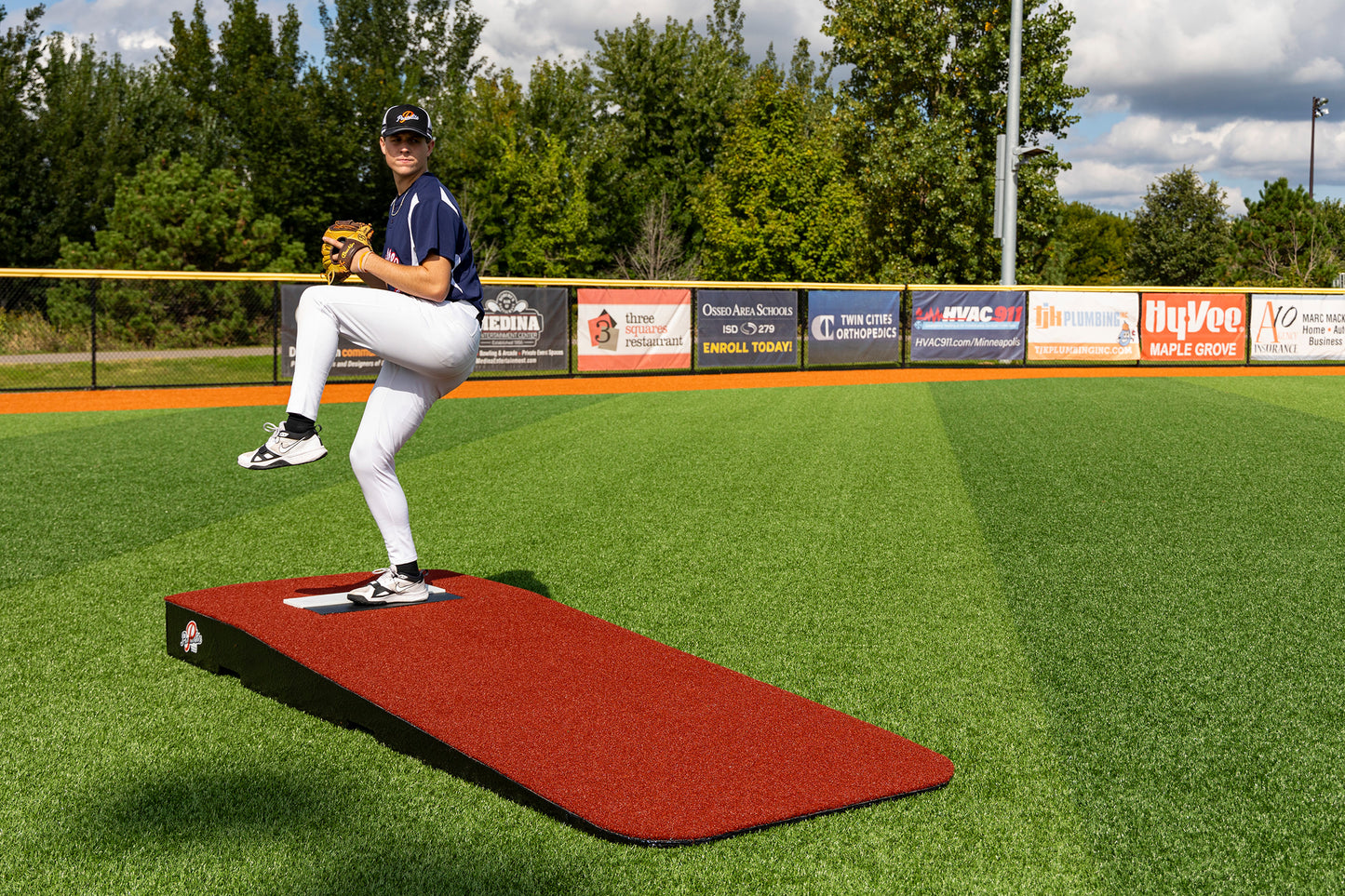 A baseball player in uniform stands mid-pitch on the Portolite Standard One-Piece Practice Mound—a durable mound for all ages—set on a green field with trees and advertisement banners in the background beneath a partly cloudy sky.