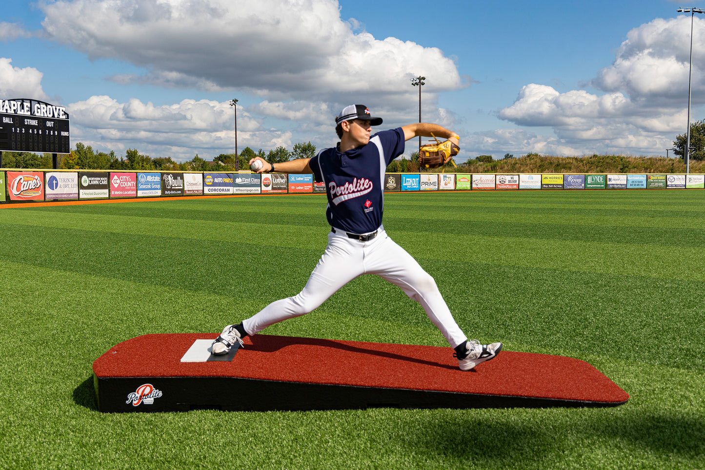 A baseball pitcher in a navy Portolite uniform winds up to throw from the Portolite Standard One-Piece Practice Mound—a durable mound for all ages—on a green turf field at Maple Grove stadium under a partly cloudy sky.