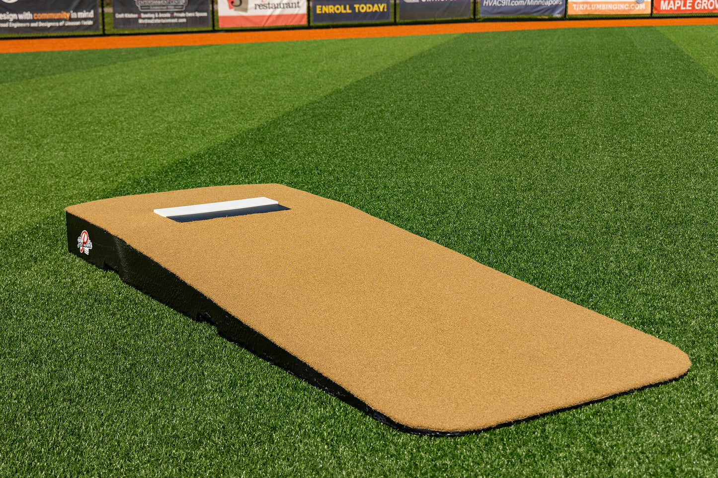 The Portolite Standard One-Piece Practice Mound, designed in a tan color, rests on vibrant green artificial turf with sports field advertisements visible in the background. This durable mound for all ages is perfect for both practices and games, making it a versatile addition to any baseball facility.