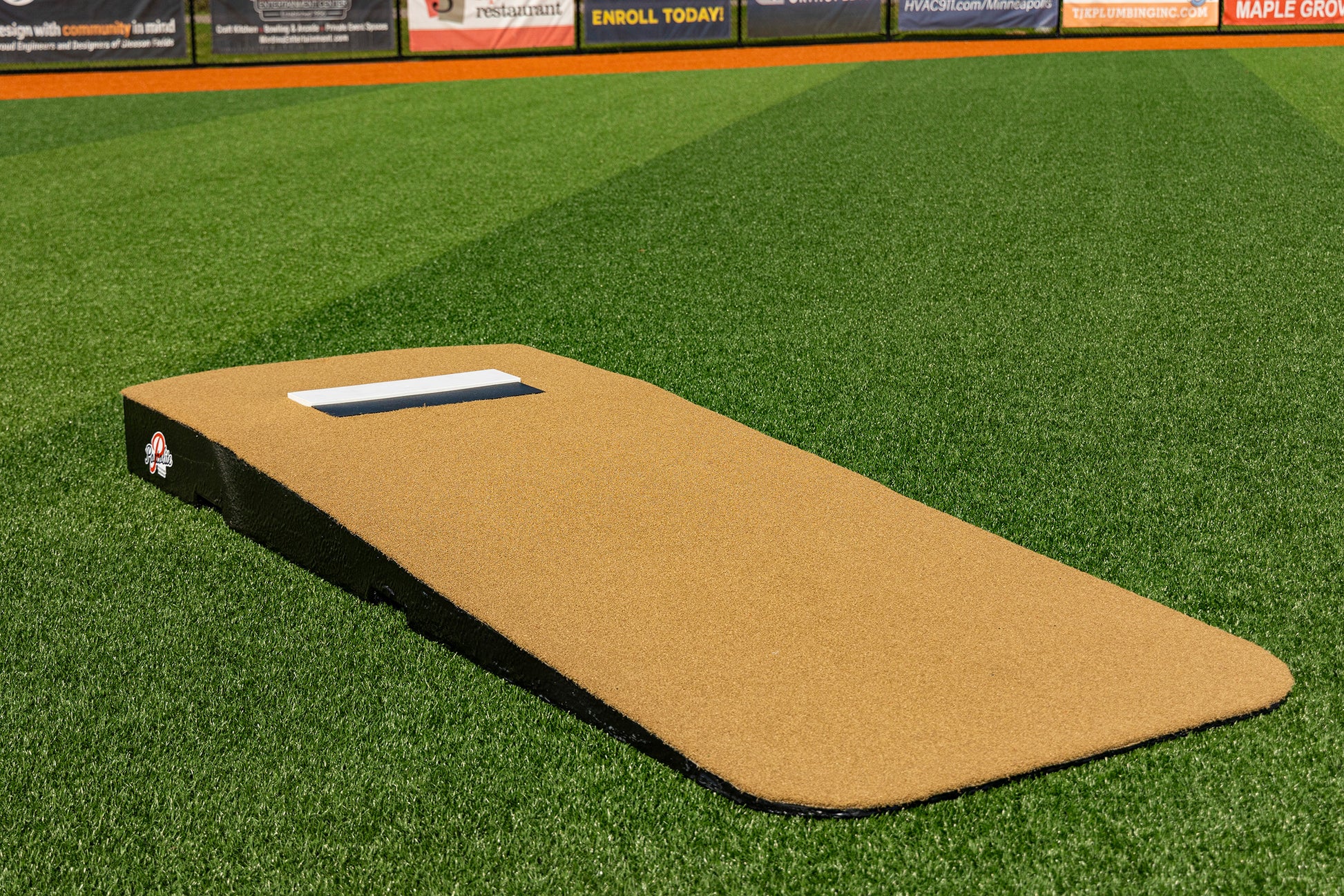 The Portolite Standard One-Piece Practice Mound, designed in a tan color, rests on vibrant green artificial turf with sports field advertisements visible in the background. This durable mound for all ages is perfect for both practices and games, making it a versatile addition to any baseball facility.