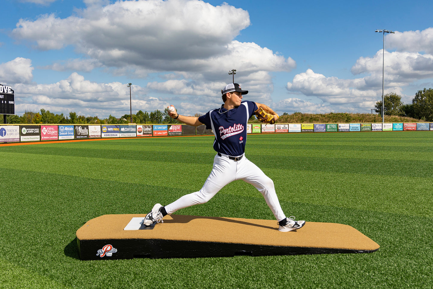 A baseball player in a navy uniform and cap is pitching on the Portolite Standard One-Piece Practice Mound, a durable mound for all ages, set on a green field under a blue sky with clouds. Advertising banners line the outfield fence.