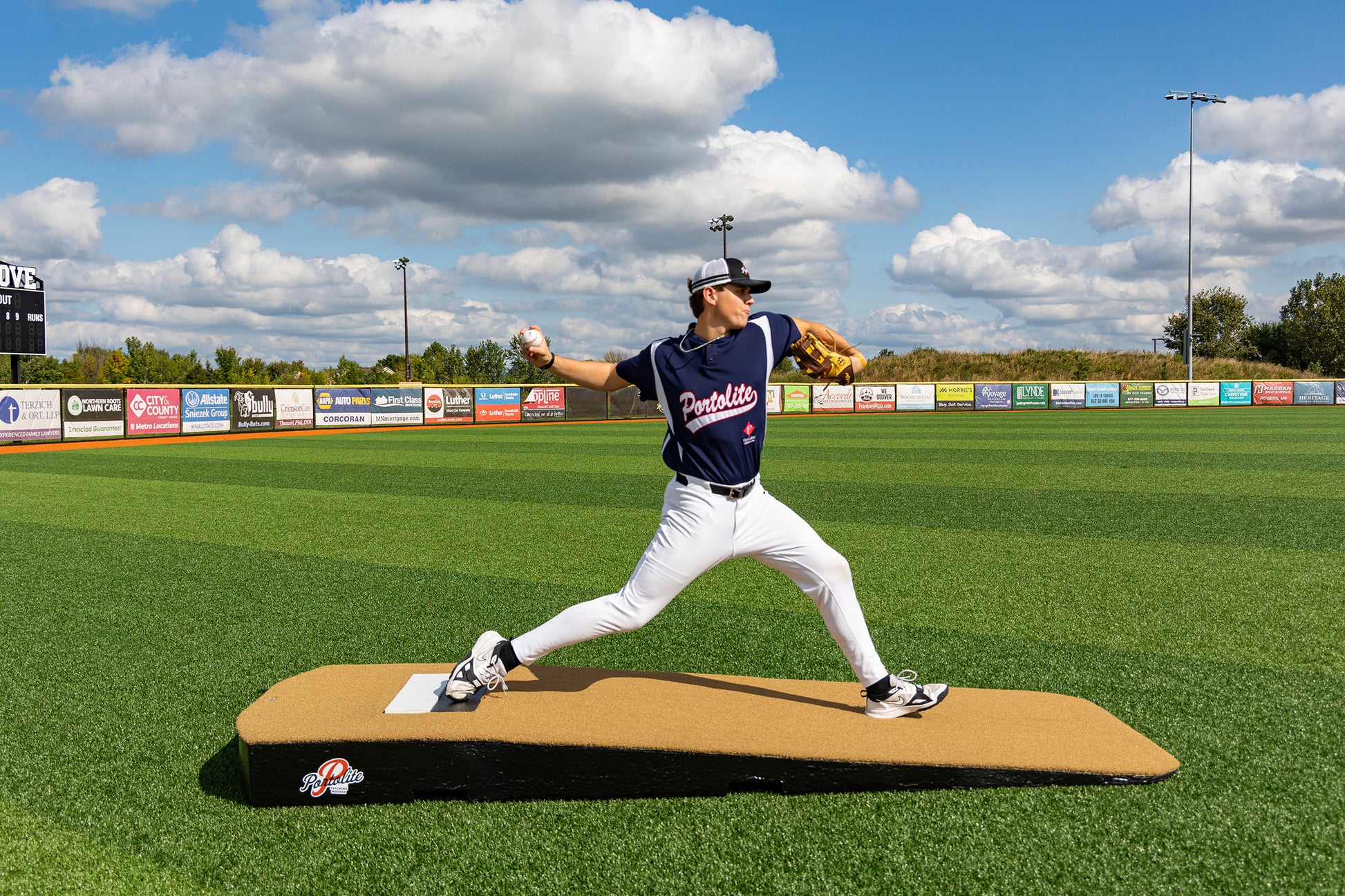 A baseball player in a navy uniform and cap is pitching on the Portolite Standard One-Piece Practice Mound, a durable mound for all ages, set on a green field under a blue sky with clouds. Advertising banners line the outfield fence.