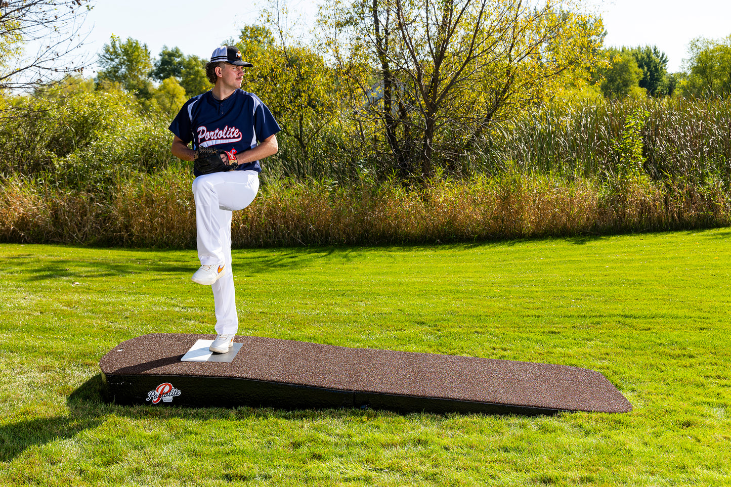 A baseball player in uniform stands on the Portolite Standard Two-Piece Practice Mound—a portable mound for all ages—outdoors, lifting one leg as if preparing to pitch, with green grass and trees in the background.