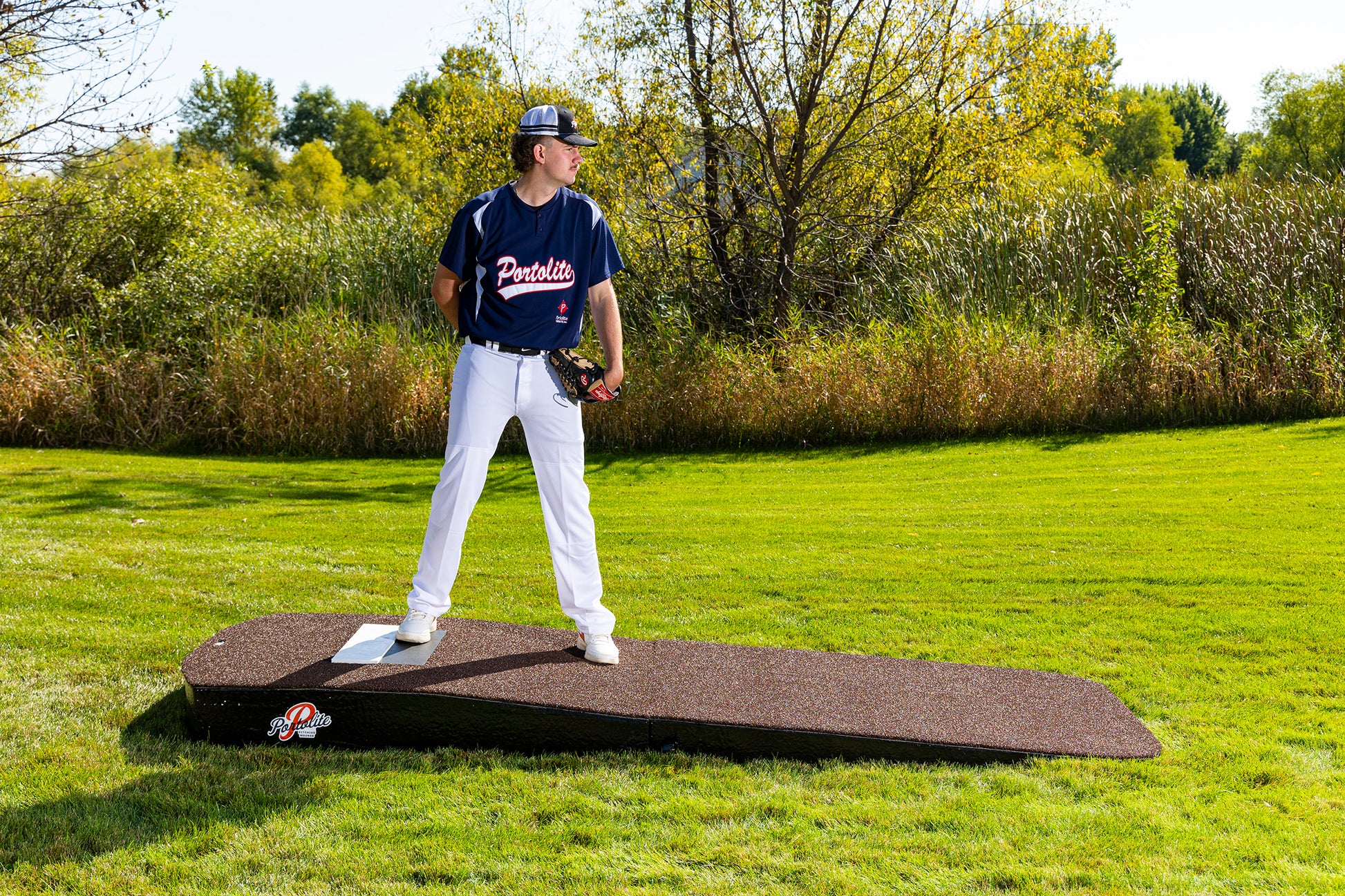 A baseball pitcher in a Portolite jersey stands on the Portolite Standard Two-Piece Practice Mound—a portable mound for all ages—outdoors on grass, holding a glove and looking toward the field, with trees in the background.