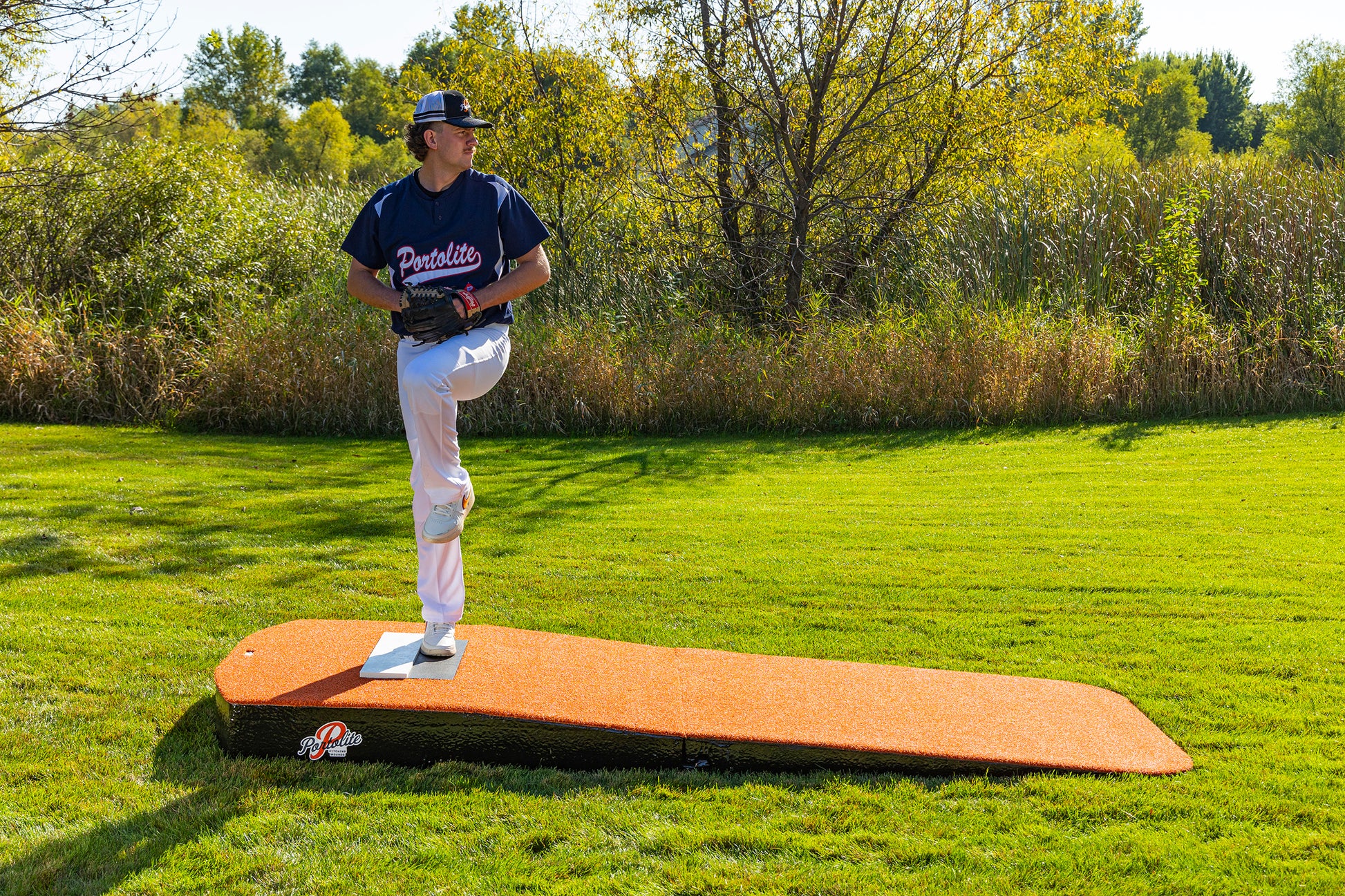 A baseball player in a blue jersey and white pants stands mid-windup on the Portolite Standard Two-Piece Practice Mound, a portable mound for all ages, set on a grassy field with trees and shrubs in the background.