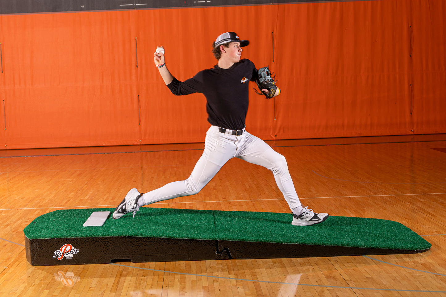 A baseball player in uniform is pitching on the Portolite Standard Two-Piece Practice Mound | Portable Mound for All Ages inside a gym with an orange wall. He has one leg forward, one leg back, and is about to throw a baseball.