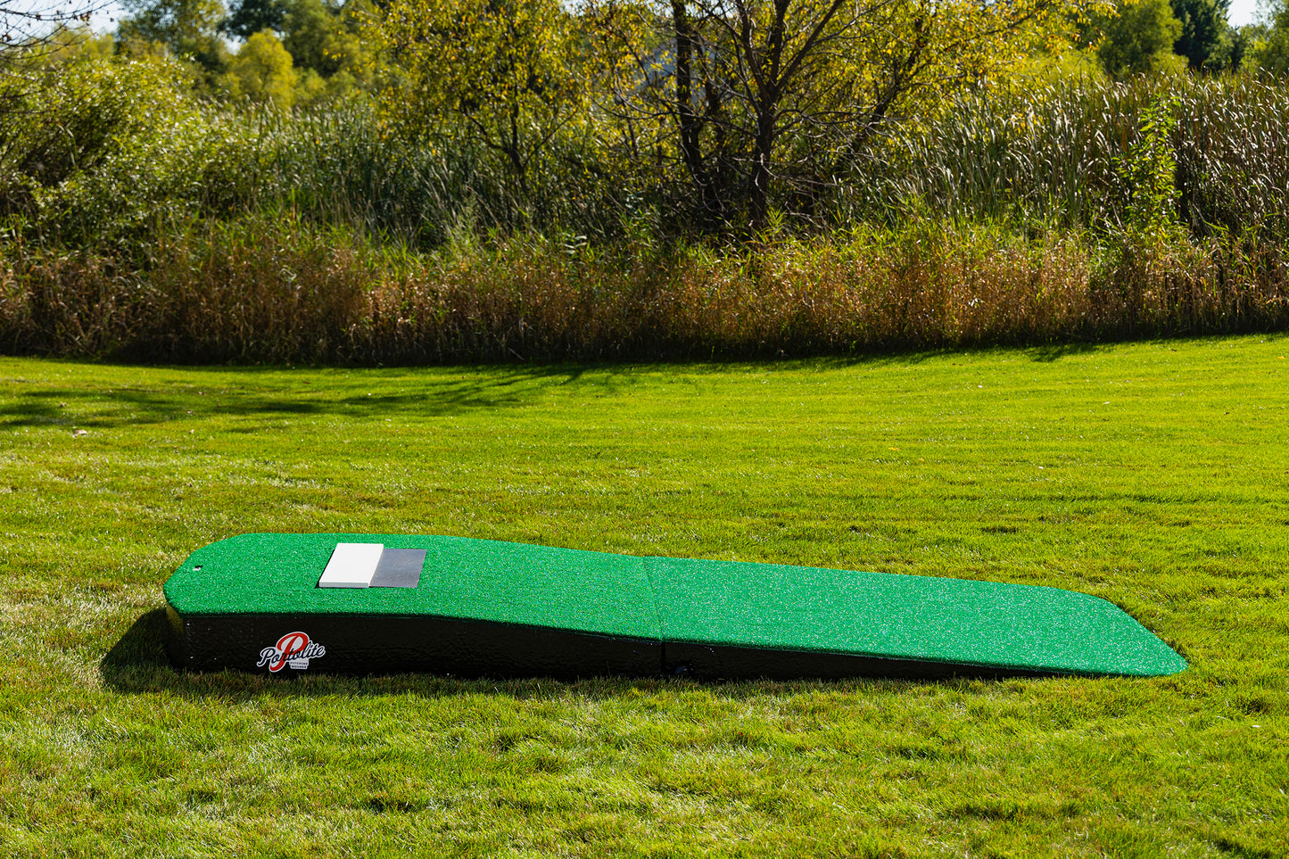 The Portolite Standard Two-Piece Practice Mound, a green, rectangular portable mound for all ages, sits on a grassy lawn with trees and tall grass in the background. Featuring an angled surface and designed to resemble a traditional pitching mound, it’s perfect for outdoor training or fun.